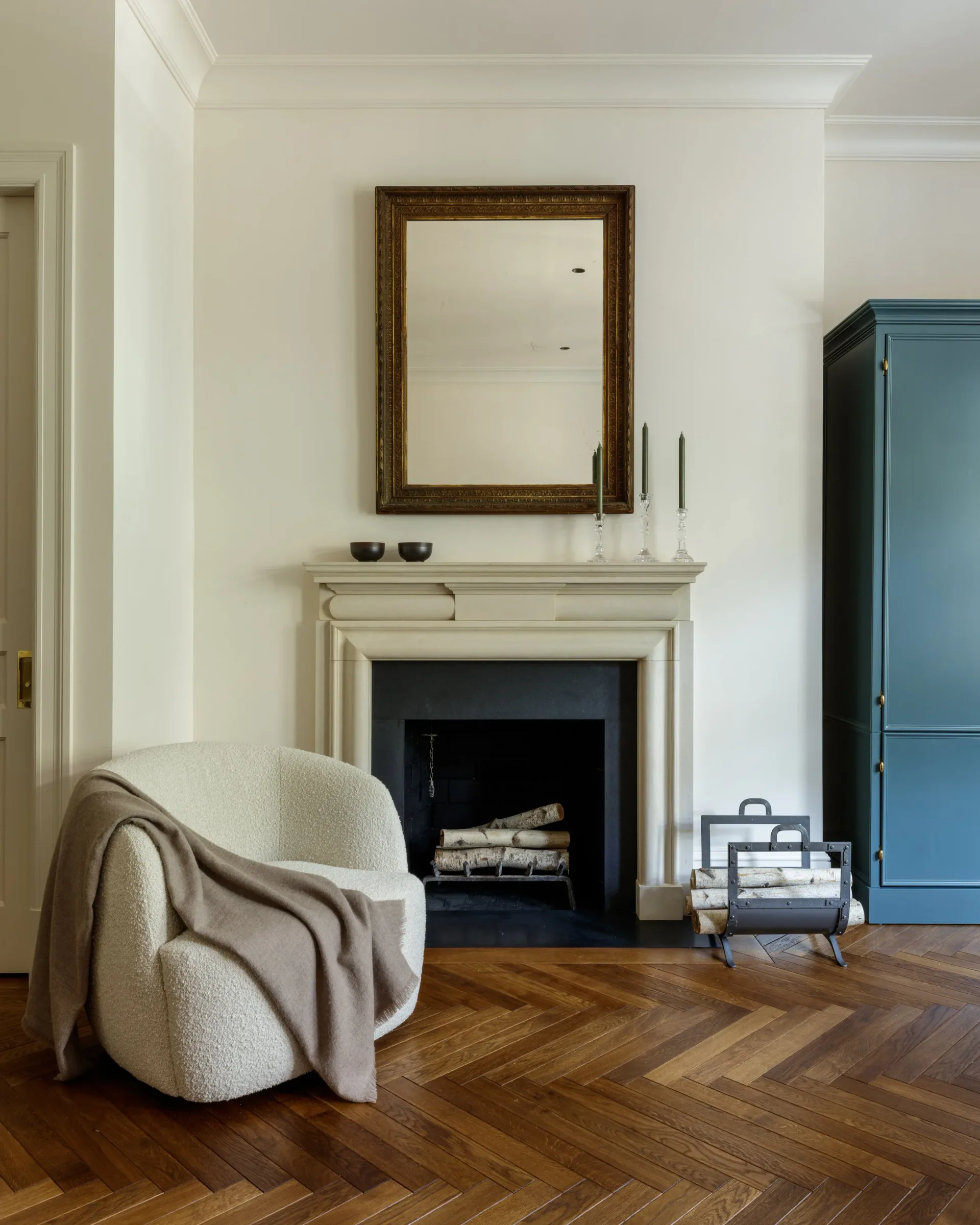 Modern fireplace living room with cream mantel, wooden herringbone flooring, curved cream armchair, and blue cabinetry by Jill Greenwood.