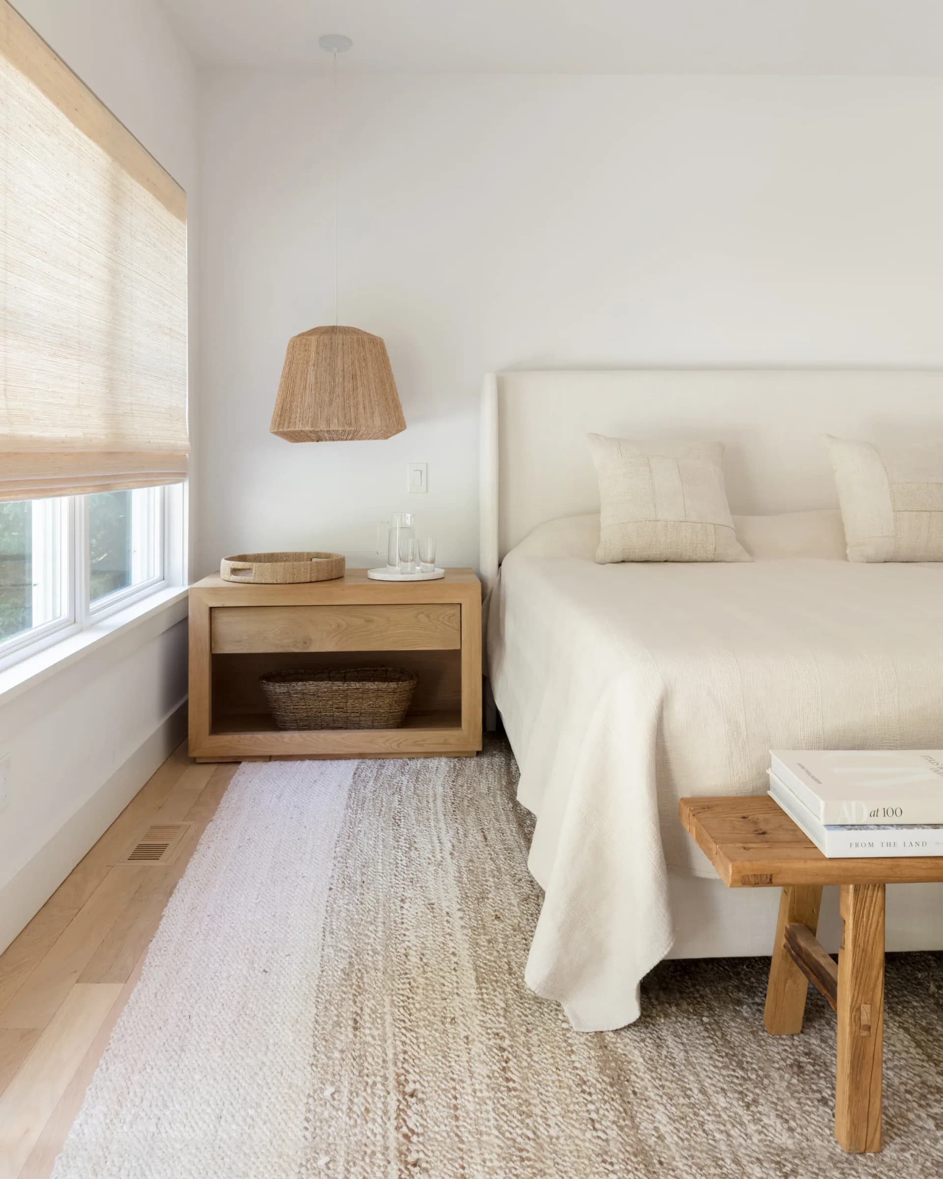 Serene bedroom with light wood nightstand, white bedding, natural linen rug, and soft natural light from window by Jill Greenwood.