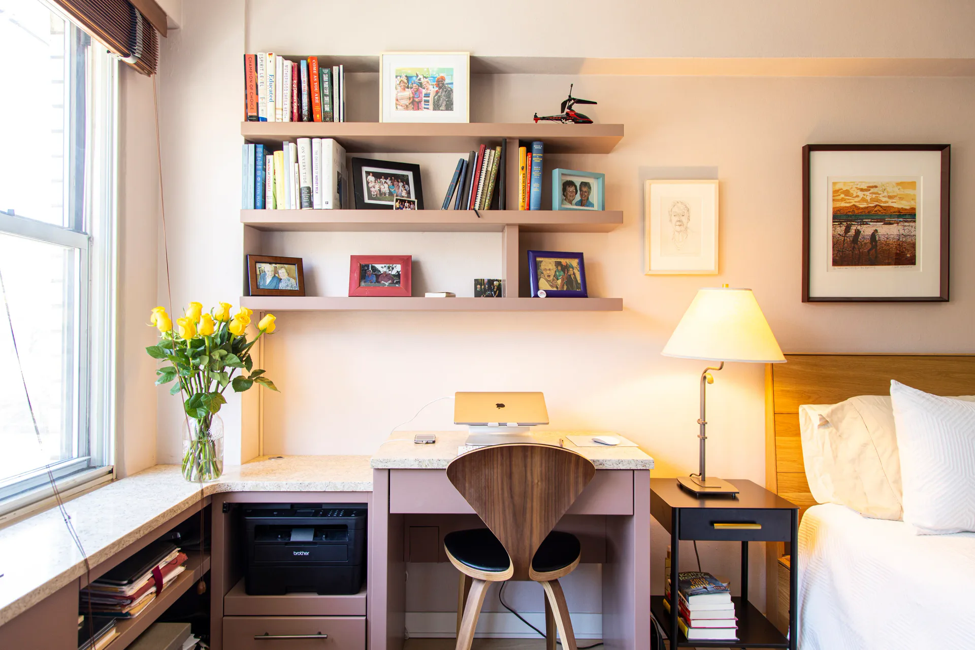 Compact bedroom workspace with floating wooden shelves, desk with wooden chair, warm lighting, and framed artwork by Jo Laurie.