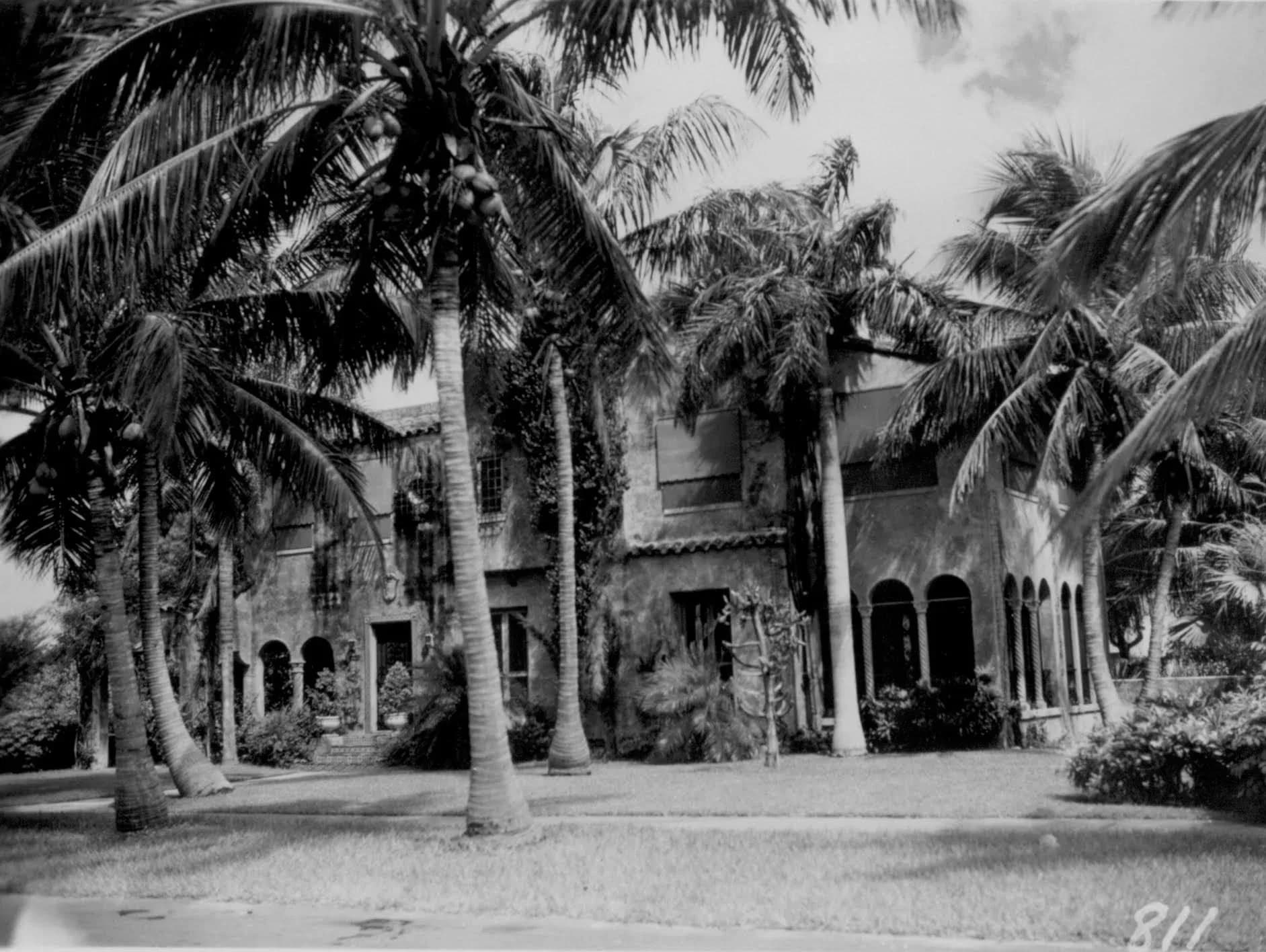 Tropical modernist villa surrounded by tall palm trees, featuring stone and stucco exterior with arched windows and openings, designed by Joanna Smith.