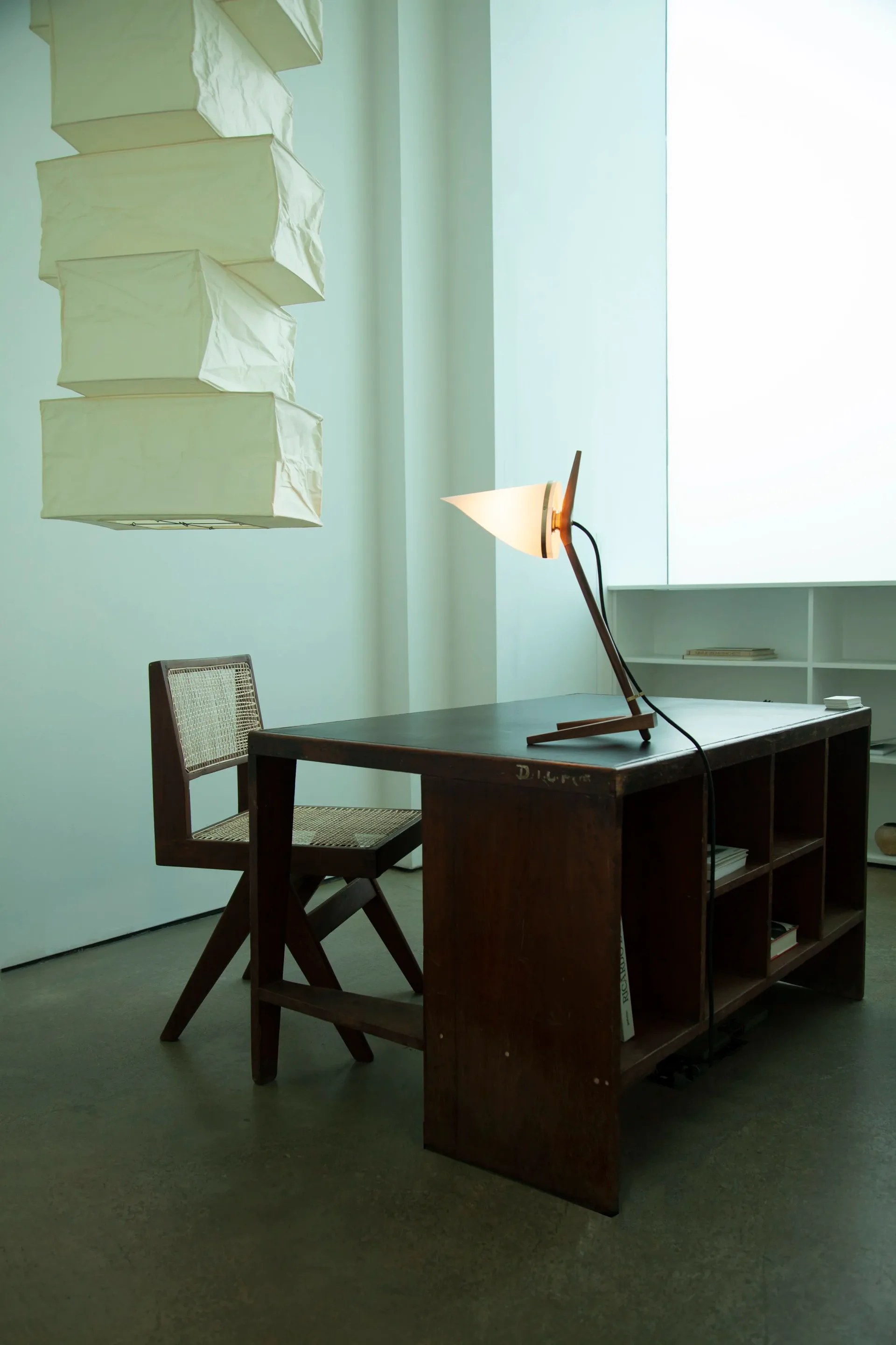 Minimalist dark wood desk with integrated shelving and matching chair, illuminated by a sculptural desk lamp, against pale green walls with stacked cream boxes overhead by Joe Armitage.