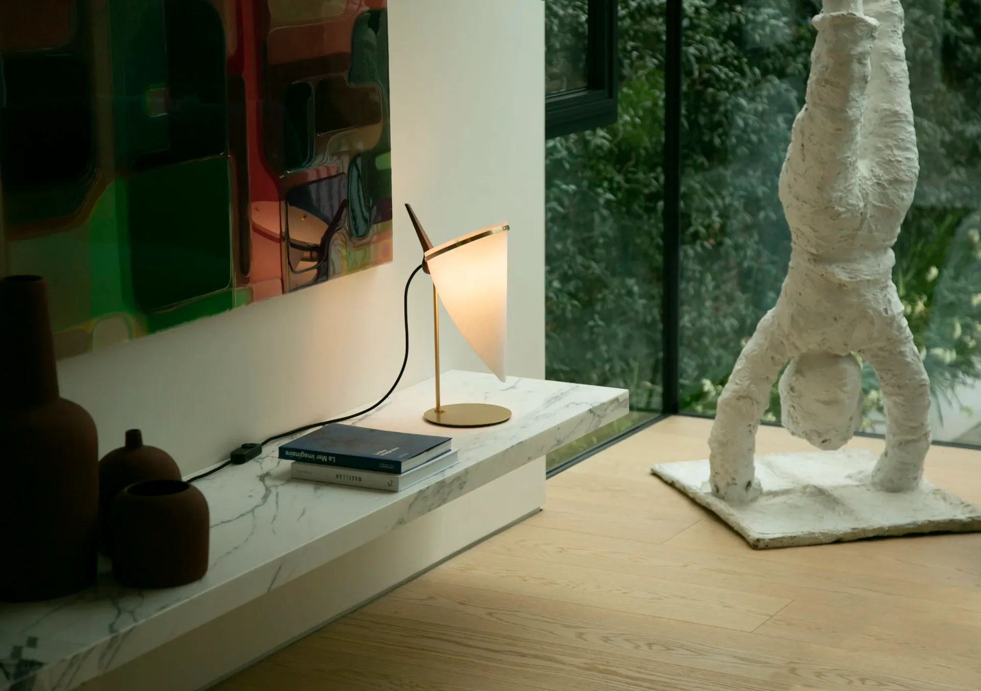 Minimalist desk workspace with marble surface, brass task lamp, and sculptural white plaster rocket form, overlooking greenery by Joe Armitage.