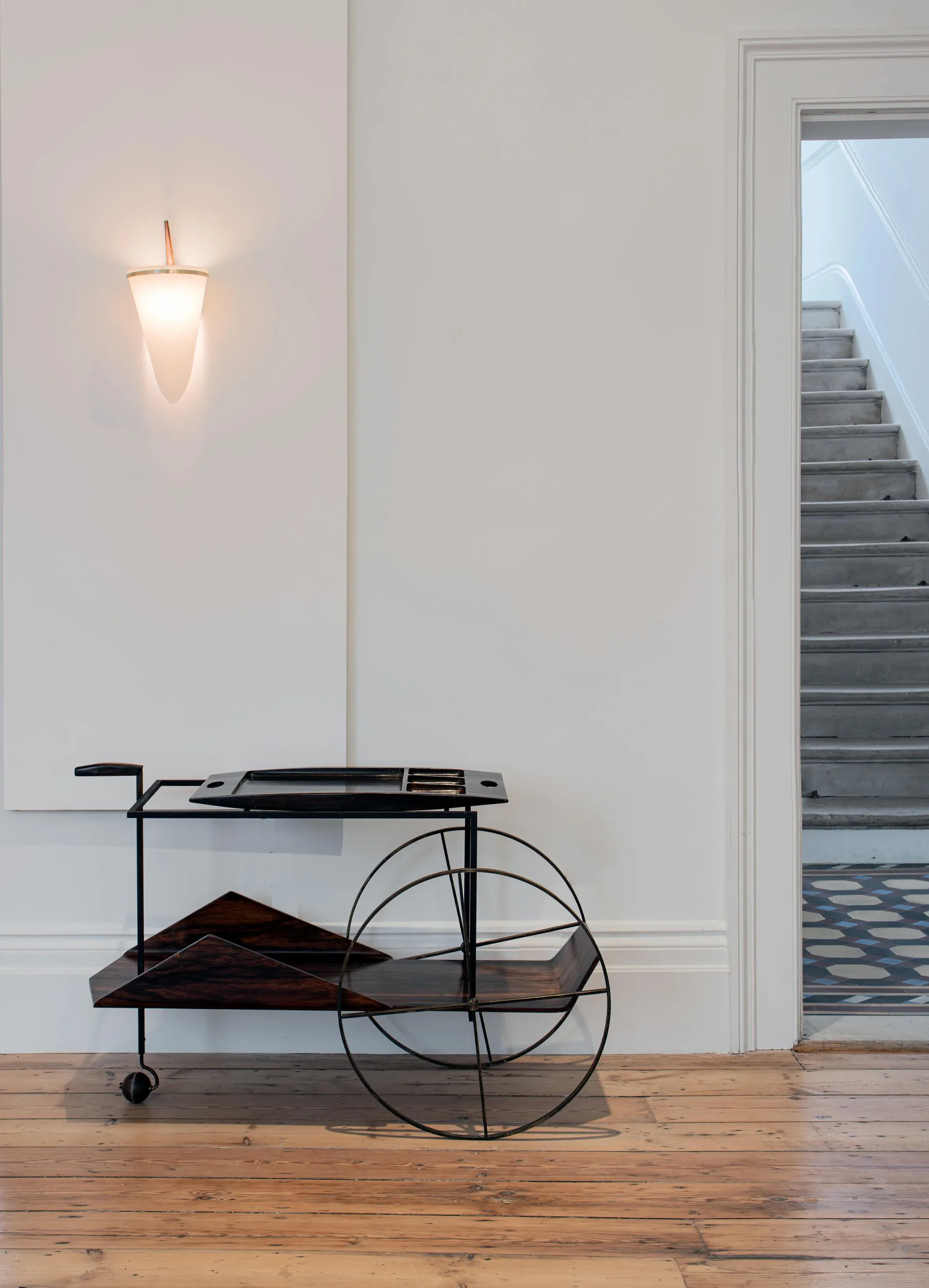 Minimalist interior with black sculptural cart on wooden floor, pendant light fixture on white wall, and staircase visible through doorway by Joe Armitage.