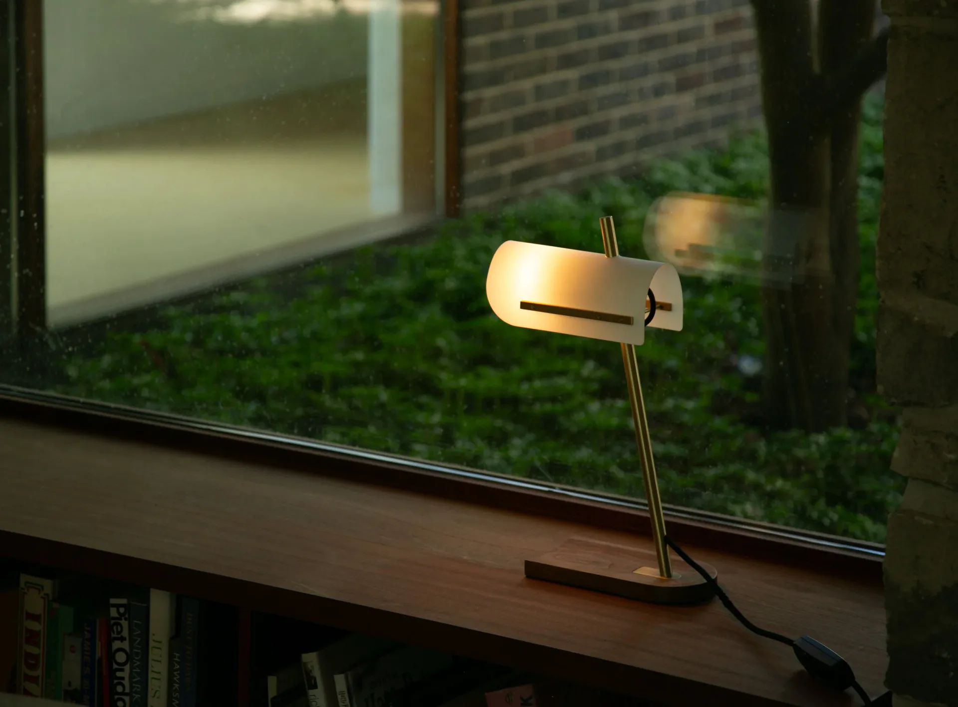 Minimalist desk lamp with warm brass finish and angular metal shade mounted on windowsill overlooking green landscape, by Joe Armitage.