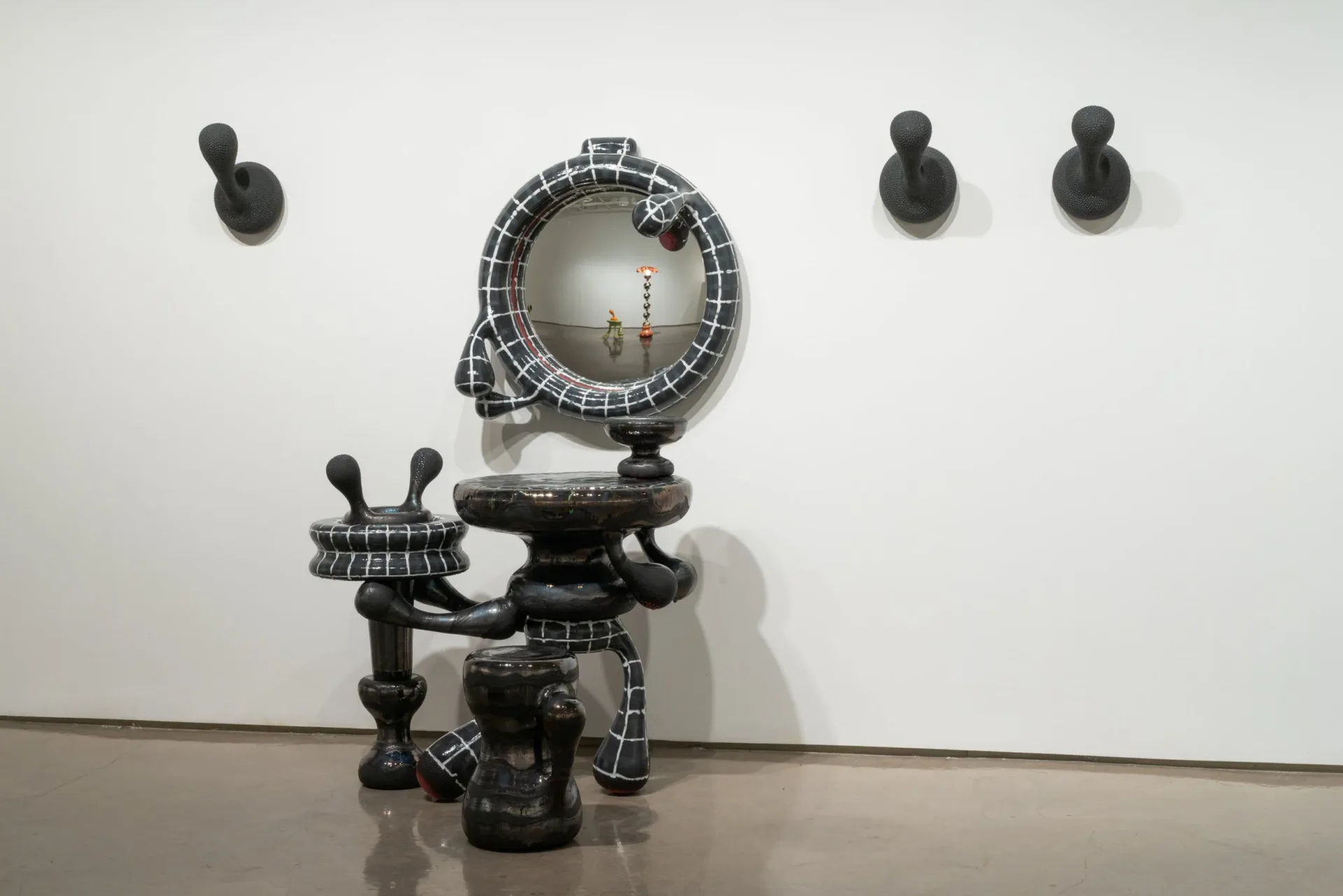 Abstract sculptural installation featuring dark geometric forms with spherical elements and a central circular mirror, mounted on white gallery wall by Joël Brodovsky-Adams.
