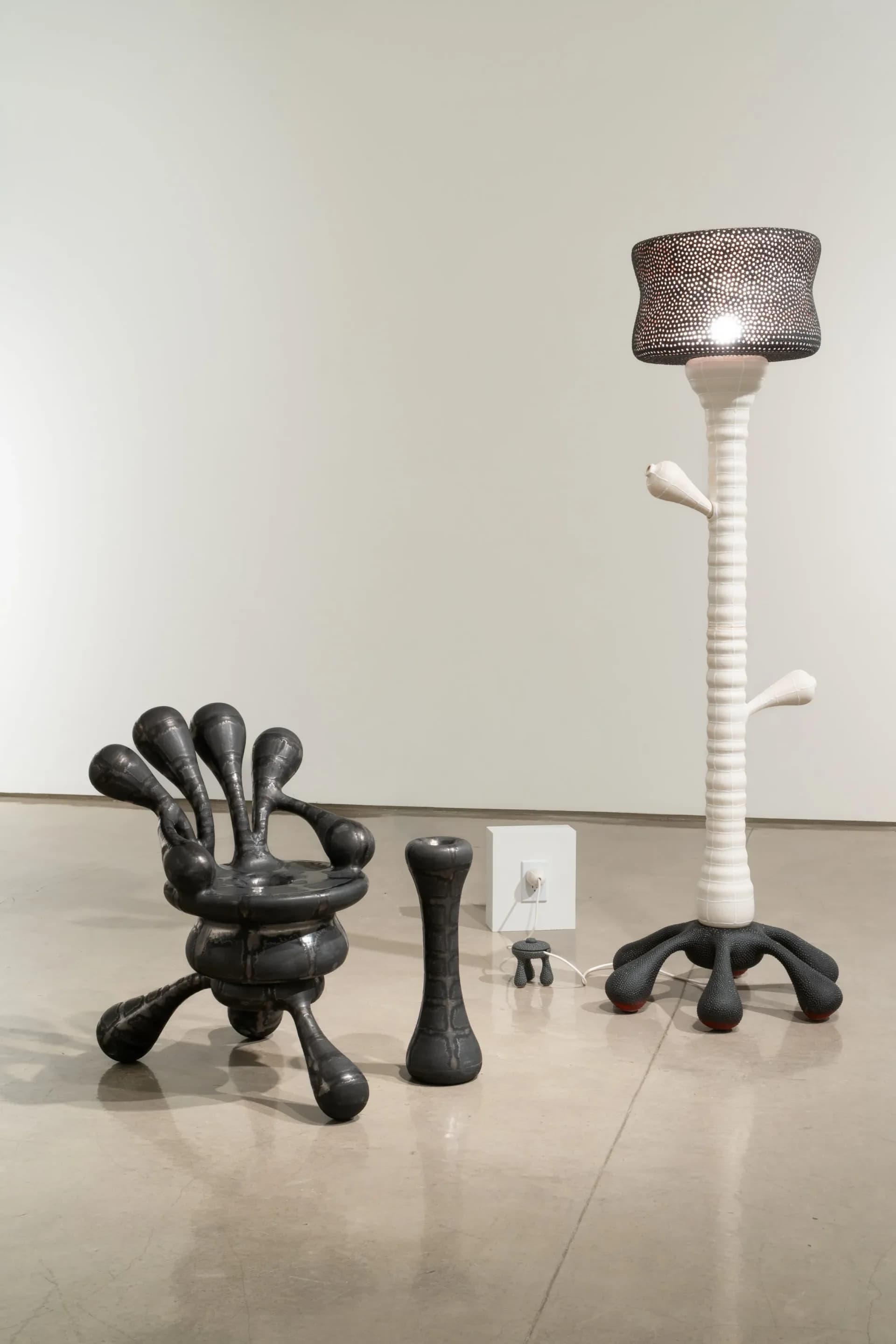 Gallery installation featuring sculptural objects in black and white, including a hand-form chair and tall stem structure with mesh head, on polished concrete floor by Joël Brodovsky-Adams.