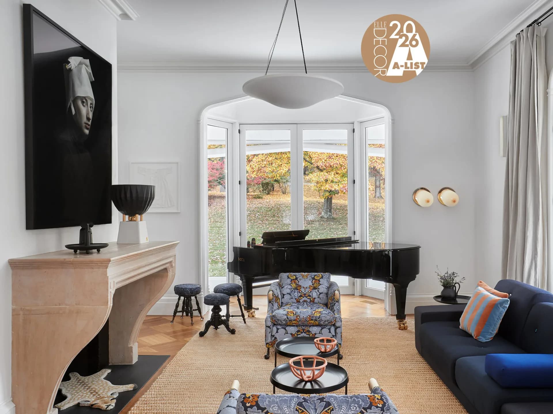 A modern living room with a grand piano, navy sofa, patterned armchair, and large multi-pane windows overlooking autumn landscape, designed by Johanna Keller.