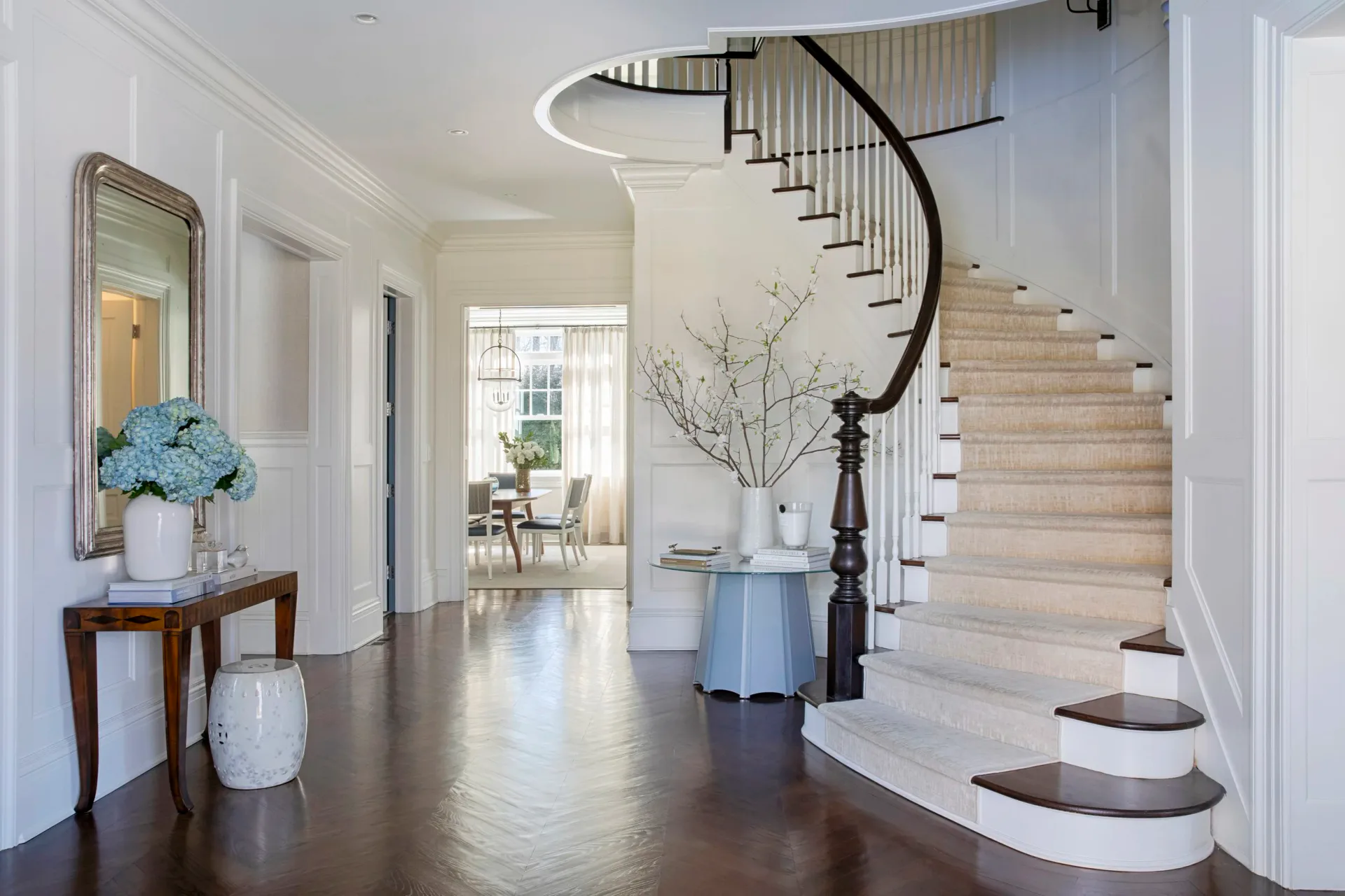 Bright foyer with curved staircase, dark wood floors, white walls, and blue hydrangea arrangement on console table by John Bjornen.