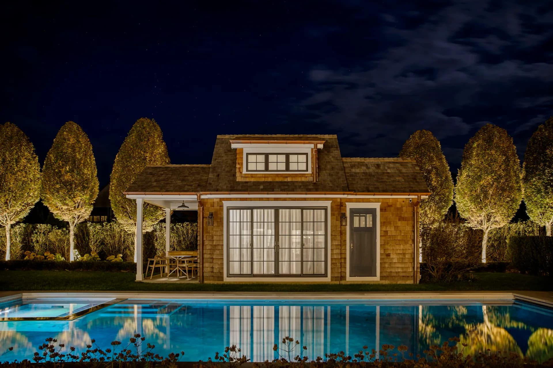 Illuminated poolhouse with warm brick exterior and large windows, reflecting in blue pool water at dusk, flanked by uplighting trees by John Bjornen.