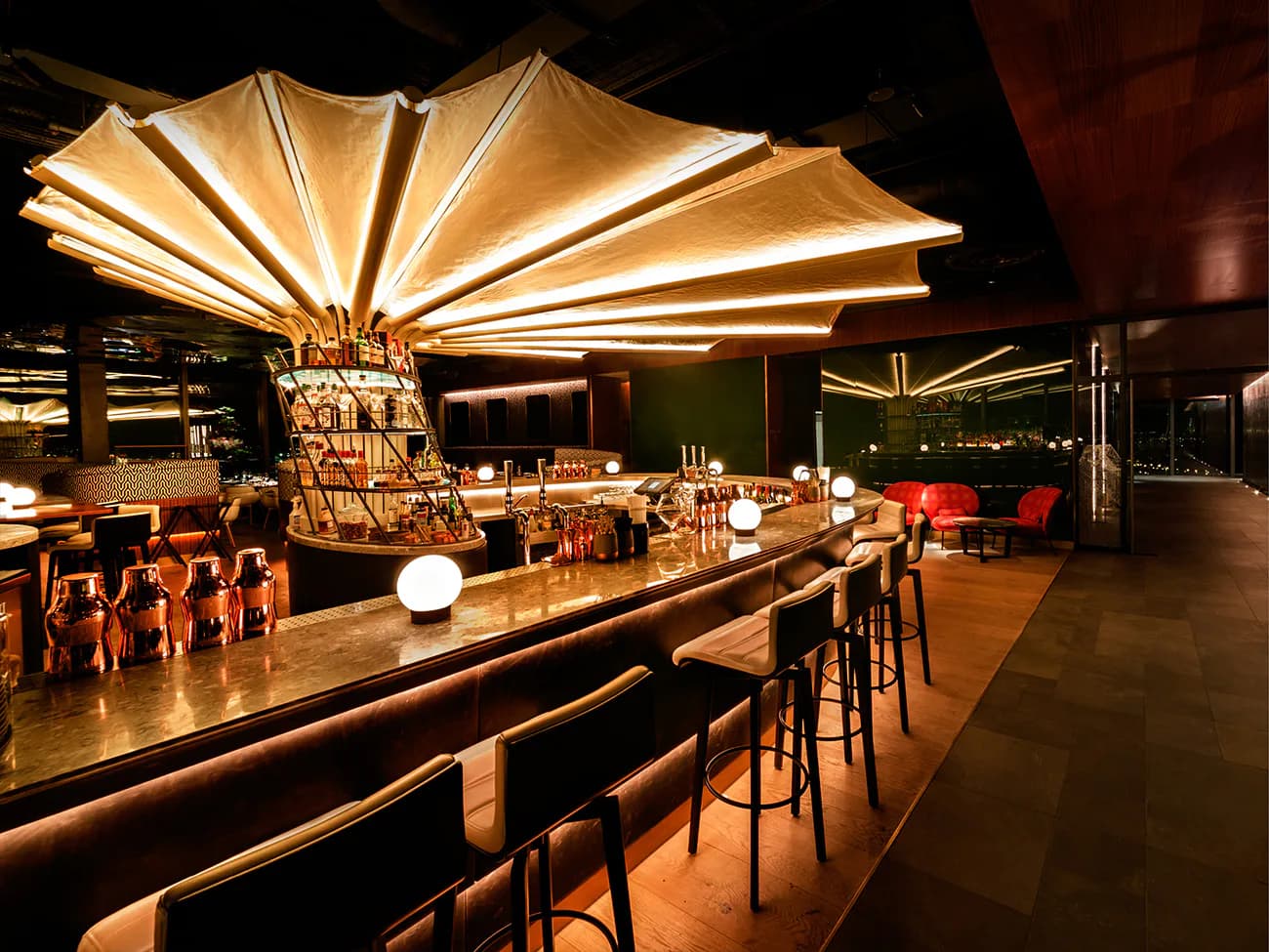 Nighttime rooftop bar with illuminated fabric canopy, wood counters, and warm ambient lighting designed by John Cetra.