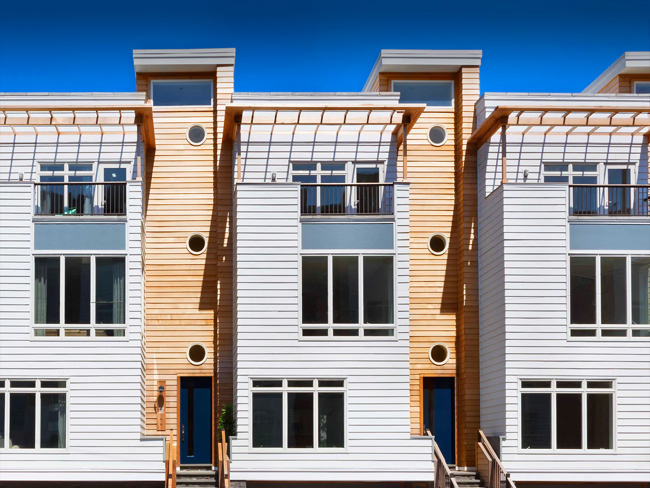 Modern residential building facade with white horizontal siding, warm wood-toned vertical pilasters, and glass balconies designed by John Cetra.