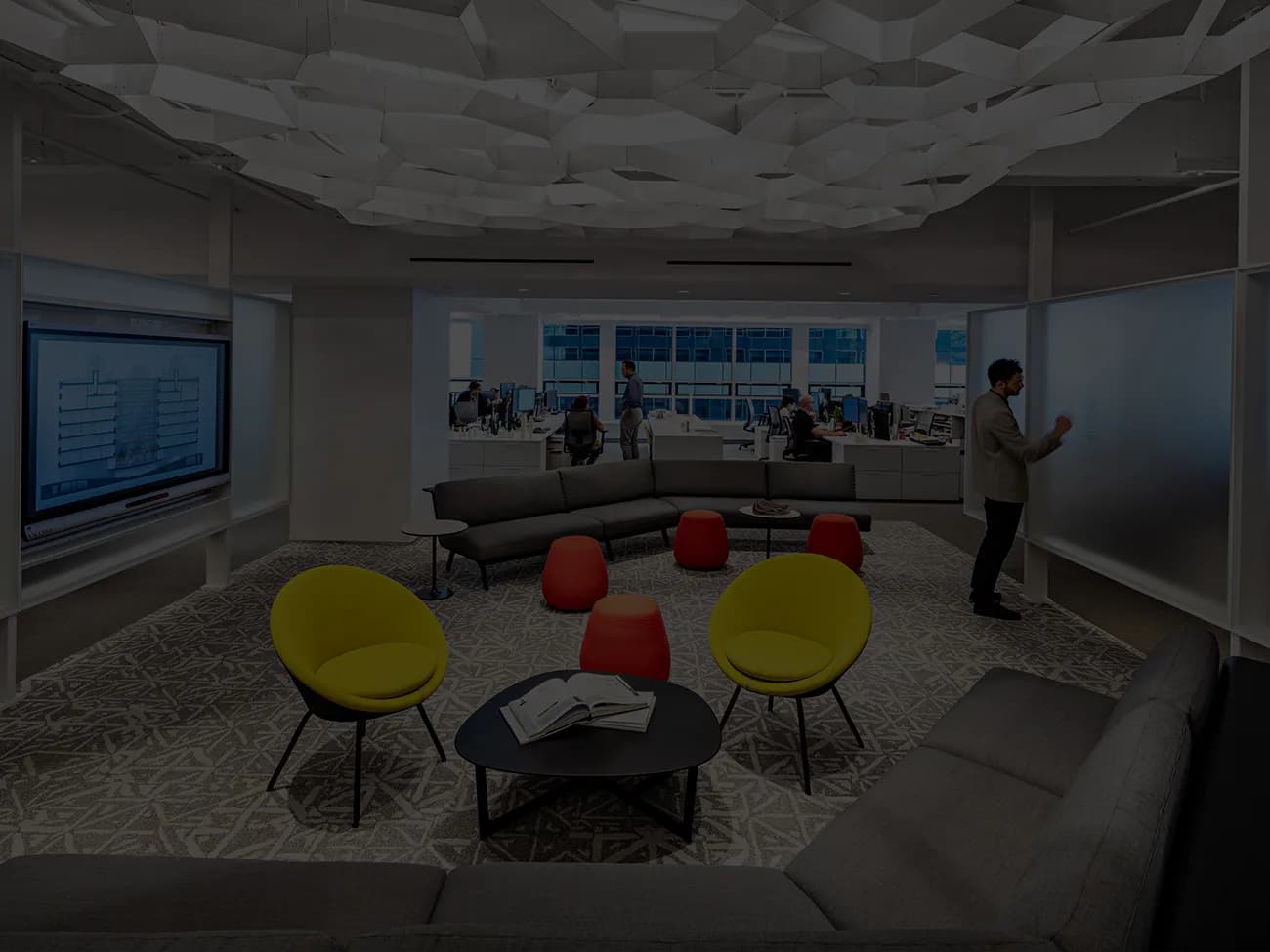 Modern office lounge with yellow chairs, red poufs, and large windows overlooking a city skyline, designed by John Cetra.