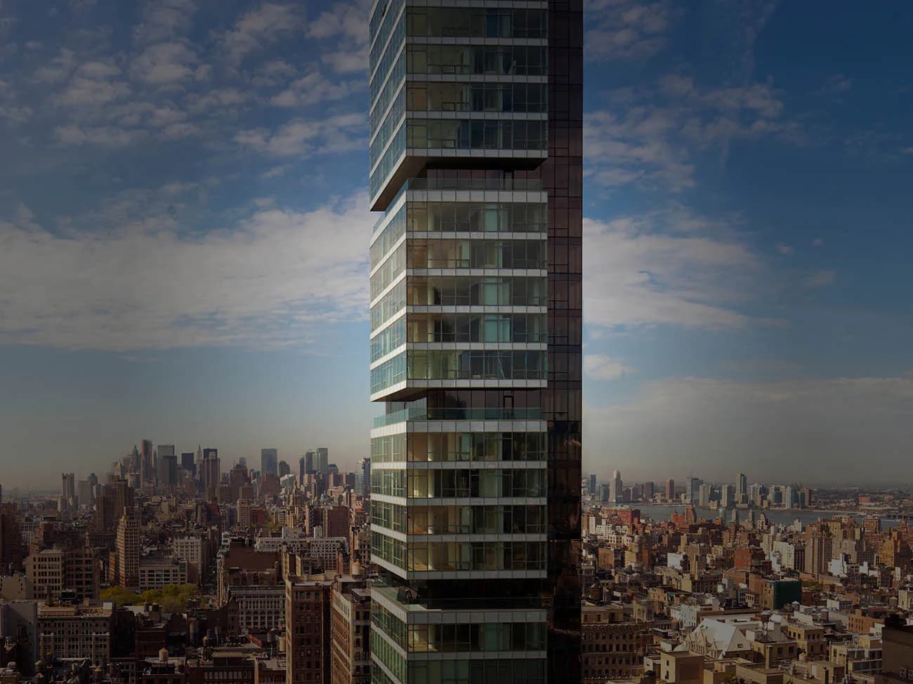 Tall residential tower with stacked glass and metal balconies overlooking Manhattan skyline by John Cetra.