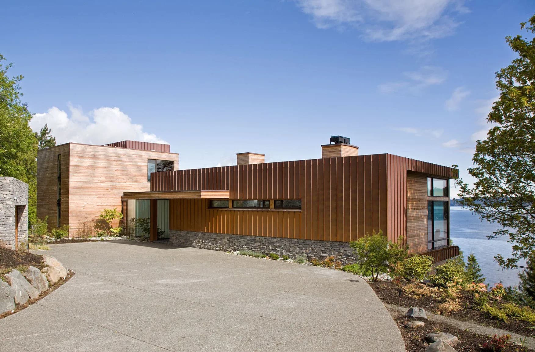 Modern residential architecture with warm wood and copper cladding overlooks water, featuring clean lines and natural landscaping by John Field.
