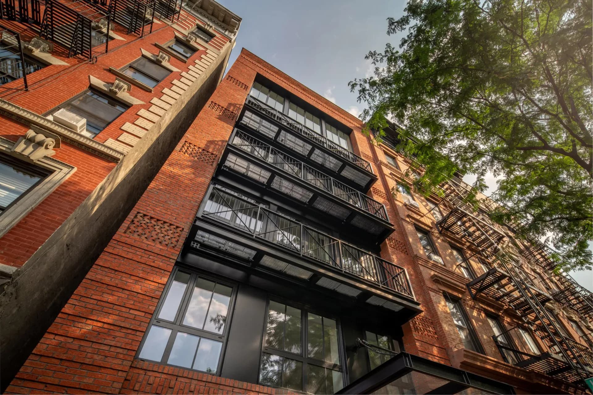 Red brick residential building facade with black metal fire escapes and large windows, framed by mature tree foliage, architectural work by John Field.