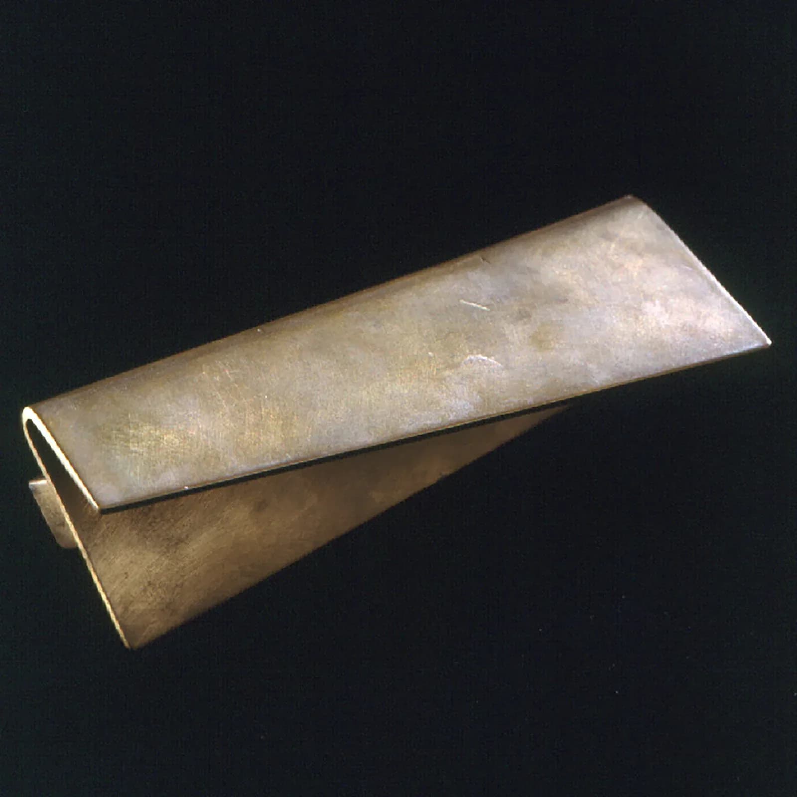 Sculptural metallic form with tapered, curved geometry in brushed brass or bronze, photographed against dark background by John Tinmouth.