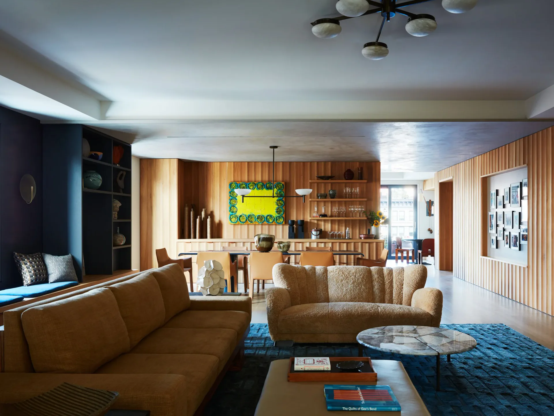 Open-plan living room with cognac leather seating, wood paneling, blue accents, and built-in shelving by Jonah Kilday.