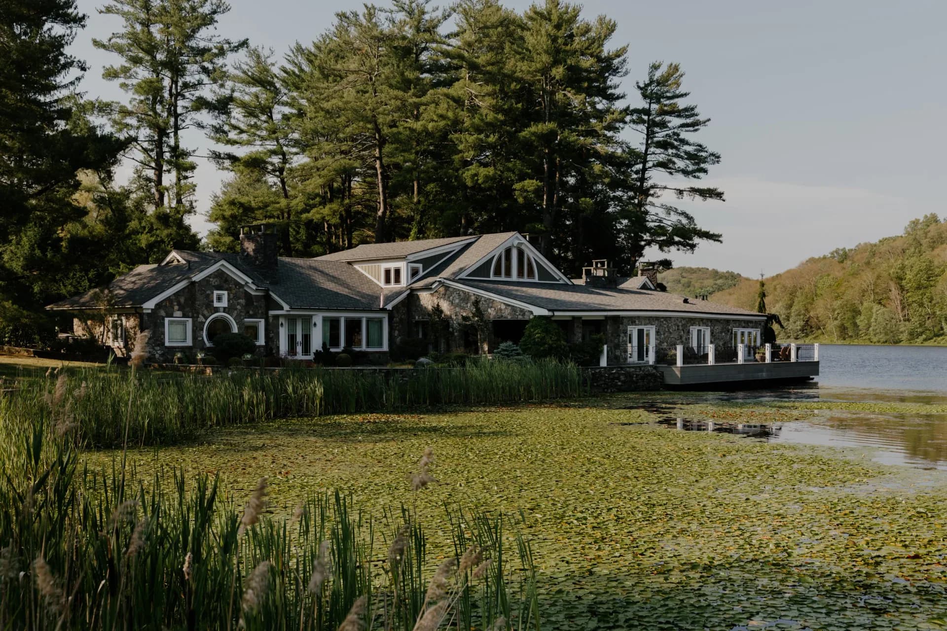 Waterfront residential complex with gray shingled boathouse and main residence set among pine trees, designed by Jonah Kilday.