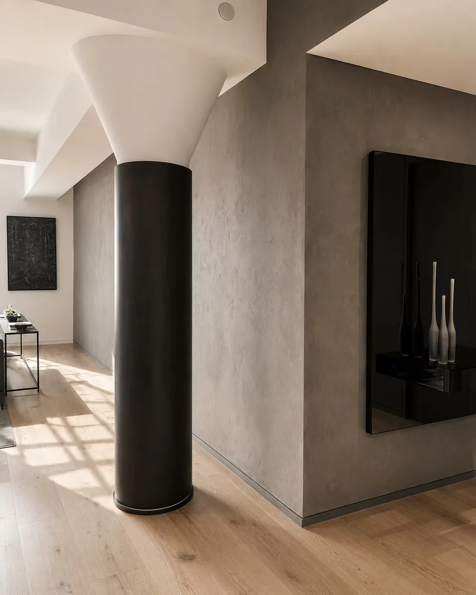 Minimalist interior with a black cylindrical column, taupe walls, white ceiling with geometric forms, and light wood flooring by Jordan Jones.