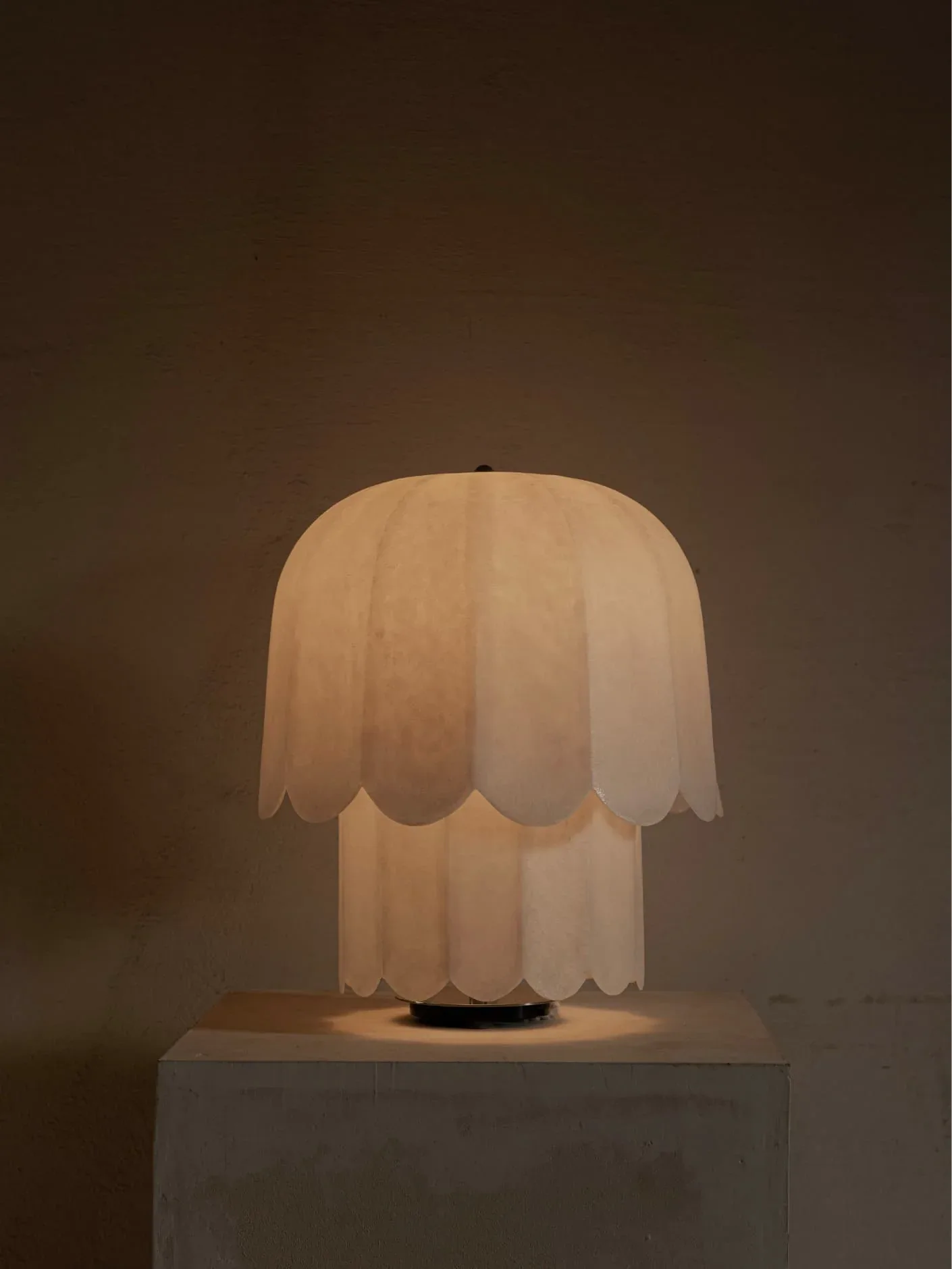 A sculptural table lamp with tiered petal-shaped white fabric shade sits illuminated on a minimalist pedestal, casting warm ambient light by Jorge Suárez-Kilzi.