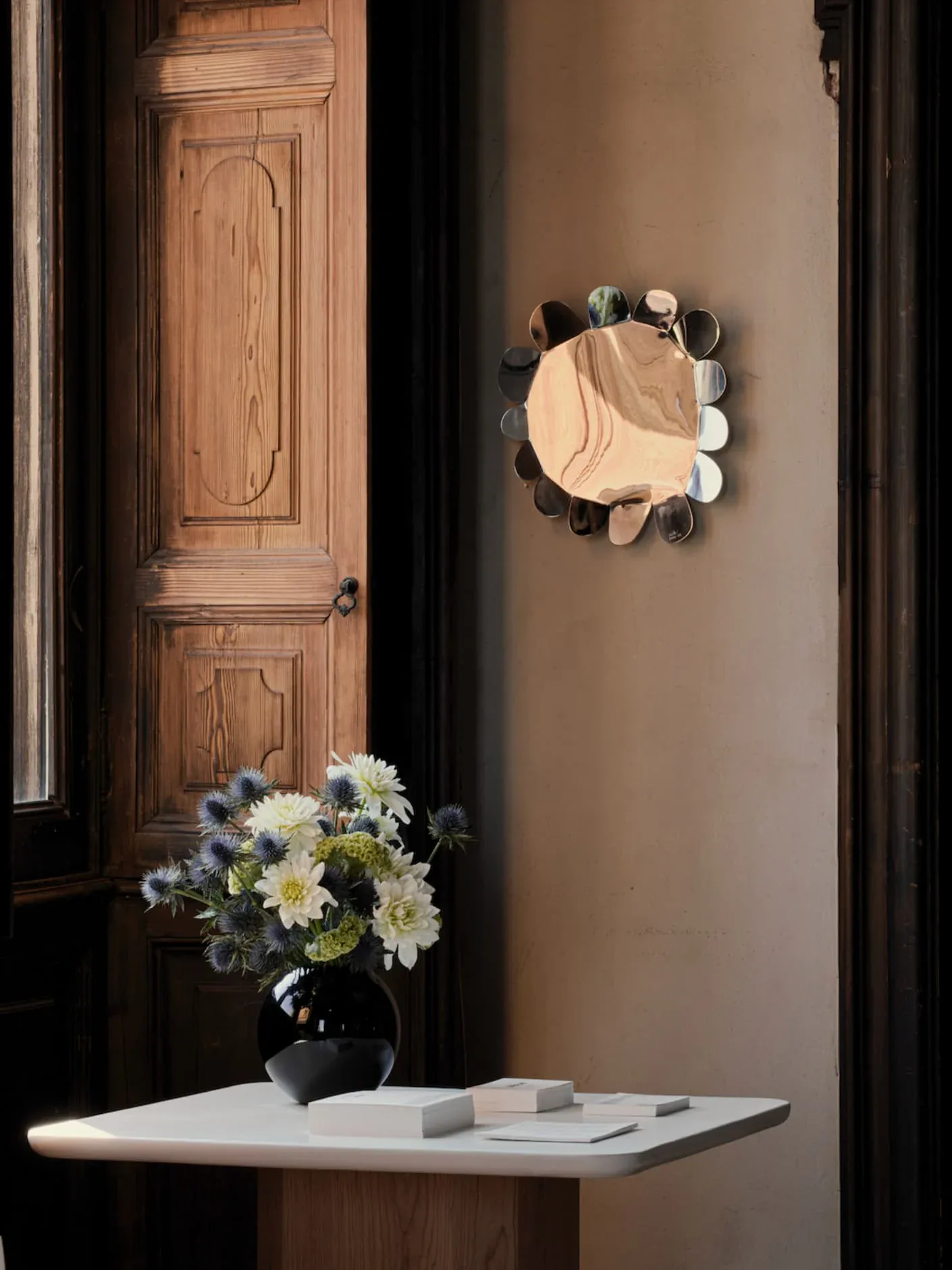 Minimalist entryway with warm taupe walls, wooden paneled door, white console table, black vase with white flowers, and circular mirror with decorative frame by Jorge Suárez-Kilzi.