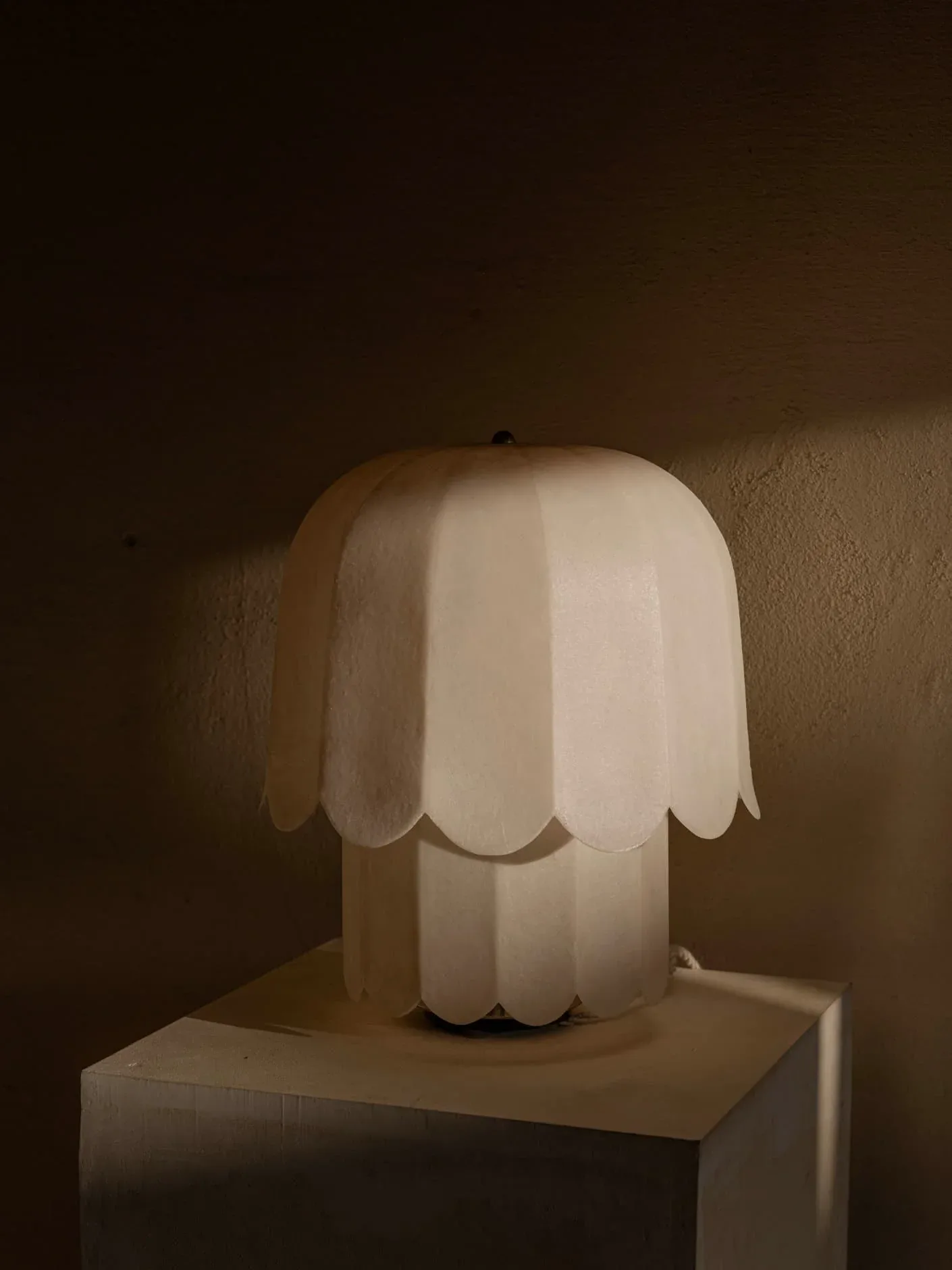 Sculptural table lamp with organic, petal-like white shade and cylindrical base on pedestal, warmly lit against neutral wall by Jorge Suárez-Kilzi.