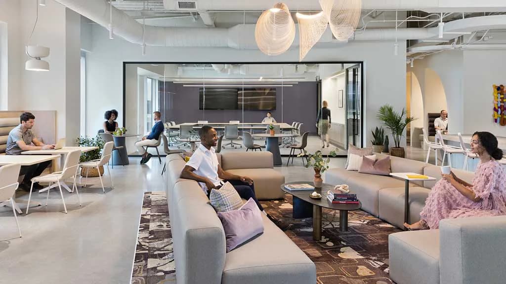 Modern office lounge with gray seating, natural wood accents, and glass-walled conference room featuring synthetic lighting and idealized spatial composition by Joseph Brancato.