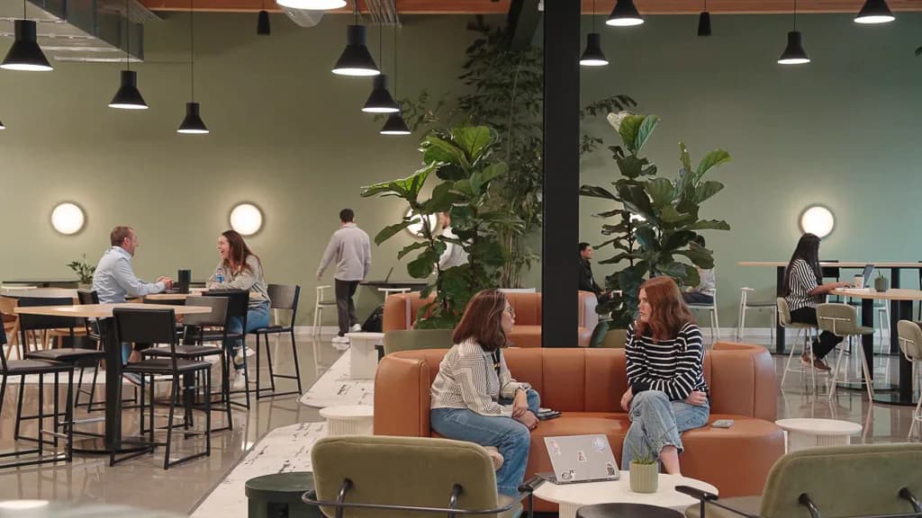 Modern office break room with terracotta seating, green walls, tall potted plants, pendant lighting, and casual workspace areas by Joseph Brancato.