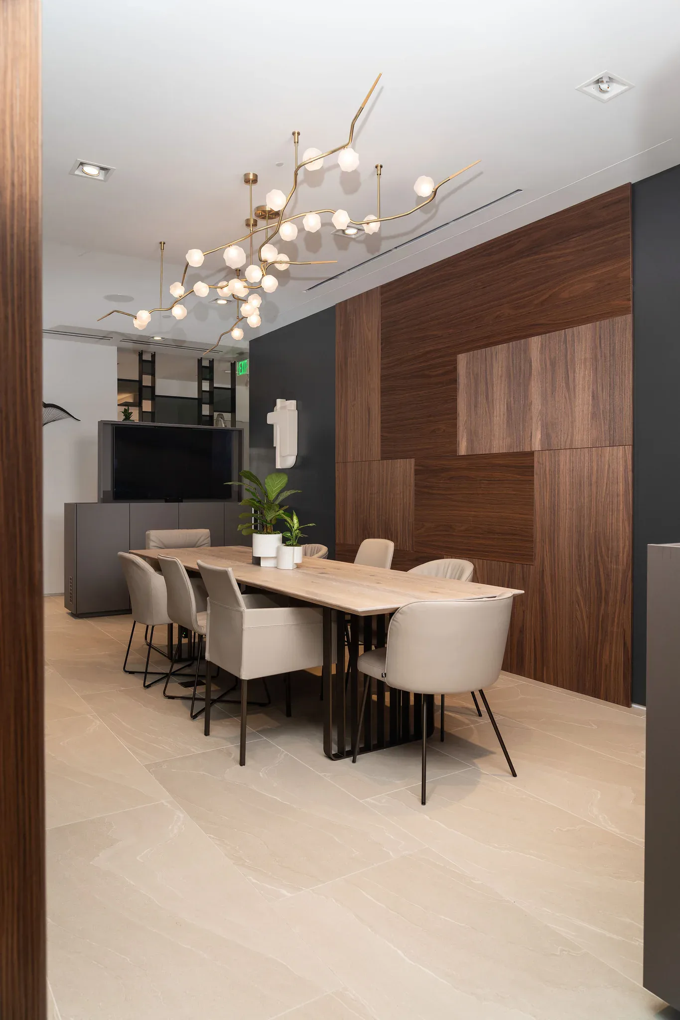 Modern dining space with wood-grain wall panels, cream chairs, natural wood table, and sculptural brass pendant lighting by Joseph Sidof.