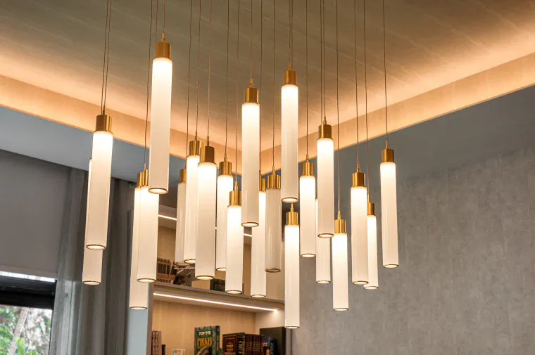 Contemporary pendant lighting installation with white cylindrical fixtures and brass accents suspended at varying heights in a residential interior by Joseph Sidof.