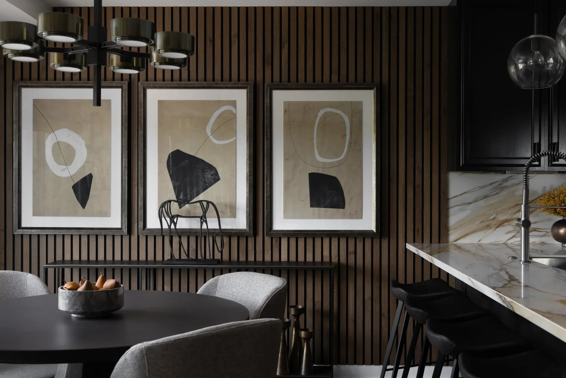 Modern dining space with vertical wood slat wall, three framed abstract artworks, black dining table, and marble kitchen island by Josh Minter.