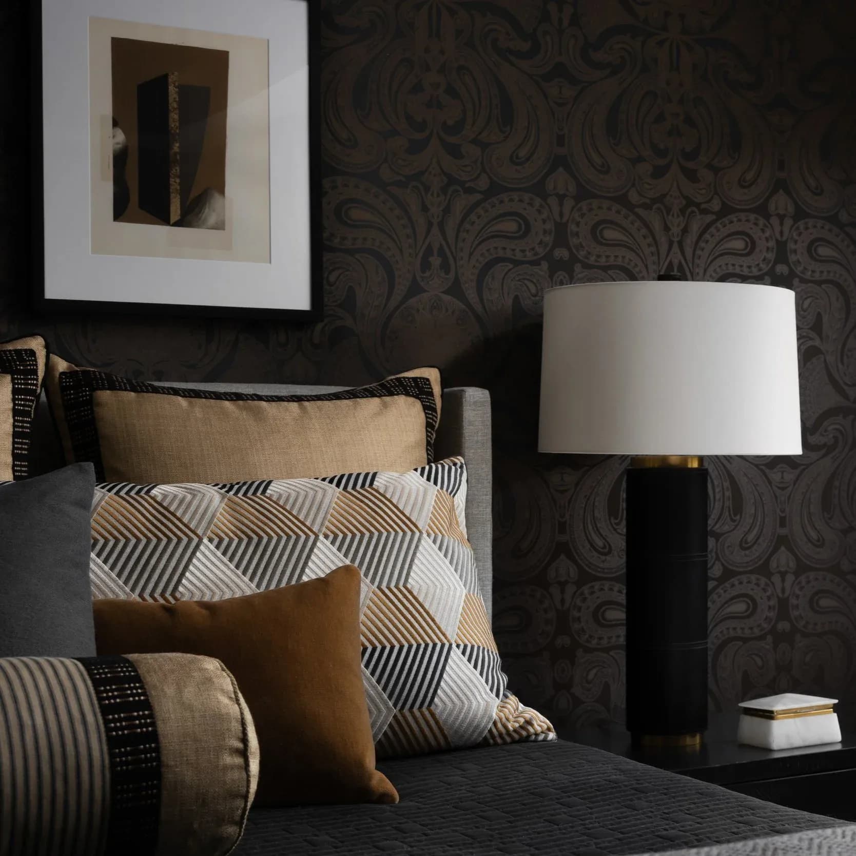 Modern bedroom with dark damask wallpaper, layered neutral bedding in tan and geometric patterns, black table lamp with white shade, and framed abstract artwork by Josh Minter.
