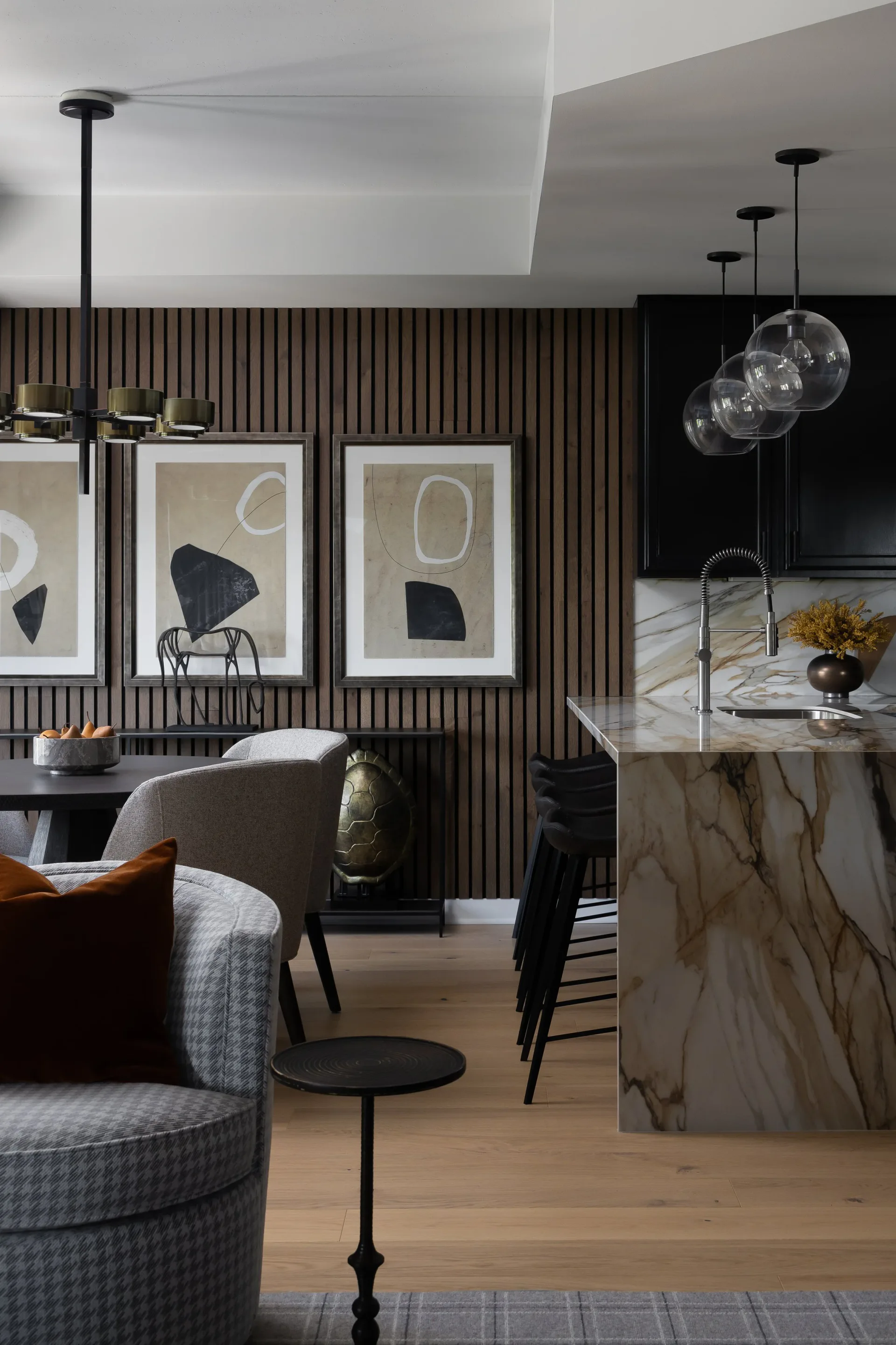Modern open-plan interior with vertical wood slat accent wall, abstract framed artwork, marble island with pendant lighting, and contemporary furnishings in neutral tones by Josh Minter.