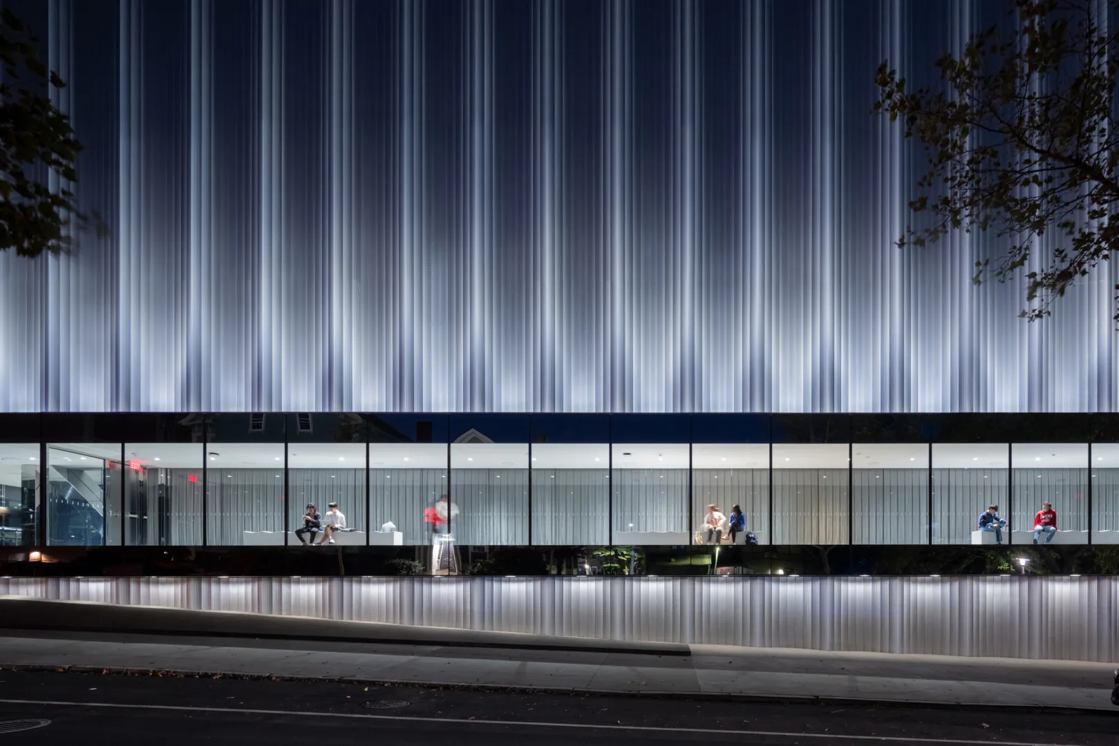 Modern glass-fronted building with illuminated corrugated metal facade and horizontal ribbon windows overlooking a street, designed by Joshua Ramus.