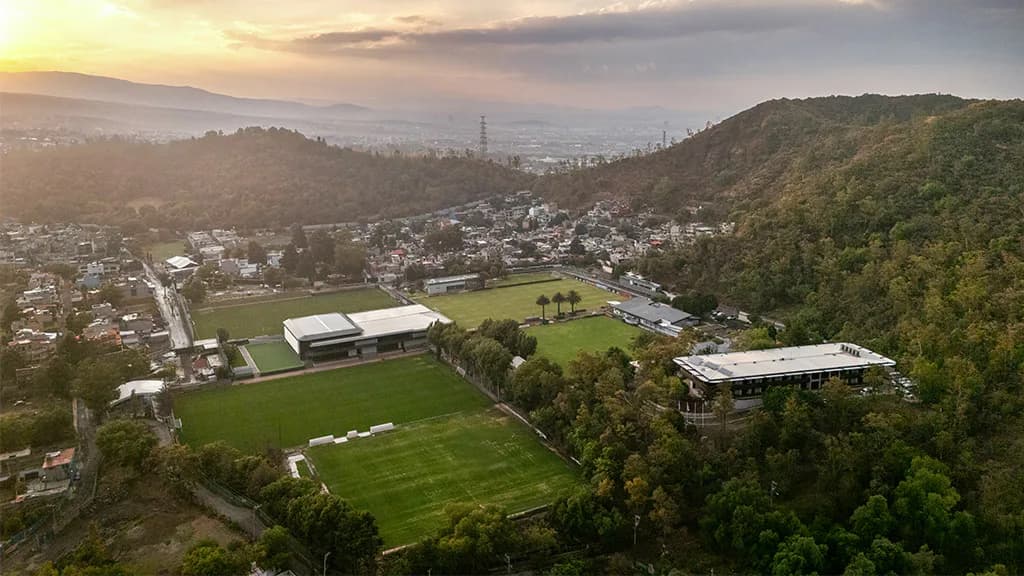 Aerial view of a modern sports facility with white structures nestled among green fields and forested hills at sunset by Ju Hyun Lee.
