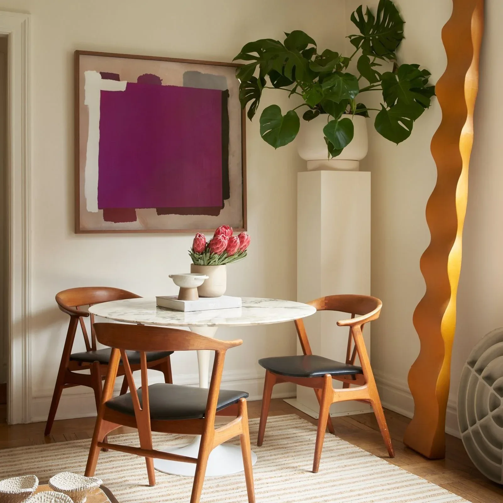 Minimalist dining room with mid-century wooden chairs, white marble table, purple abstract wall art, potted plant, and golden sculptural element by Juan J. Carretero.