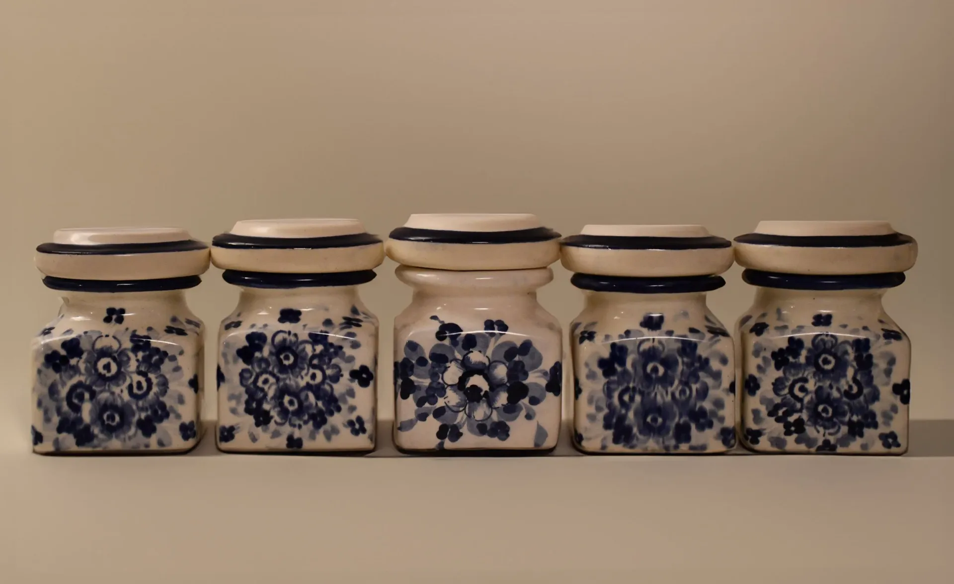 Five ceramic storage jars with blue floral patterns and dark ceramic lids arranged in a row against a neutral beige background by Juan Russo.