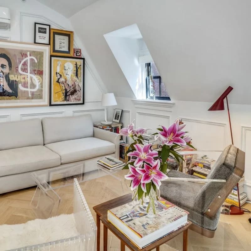 Bright attic living room with cream sectional sofa, glass coffee table, pink lilies, framed artwork, and skylight window, designed by Juanita Jimenez.
