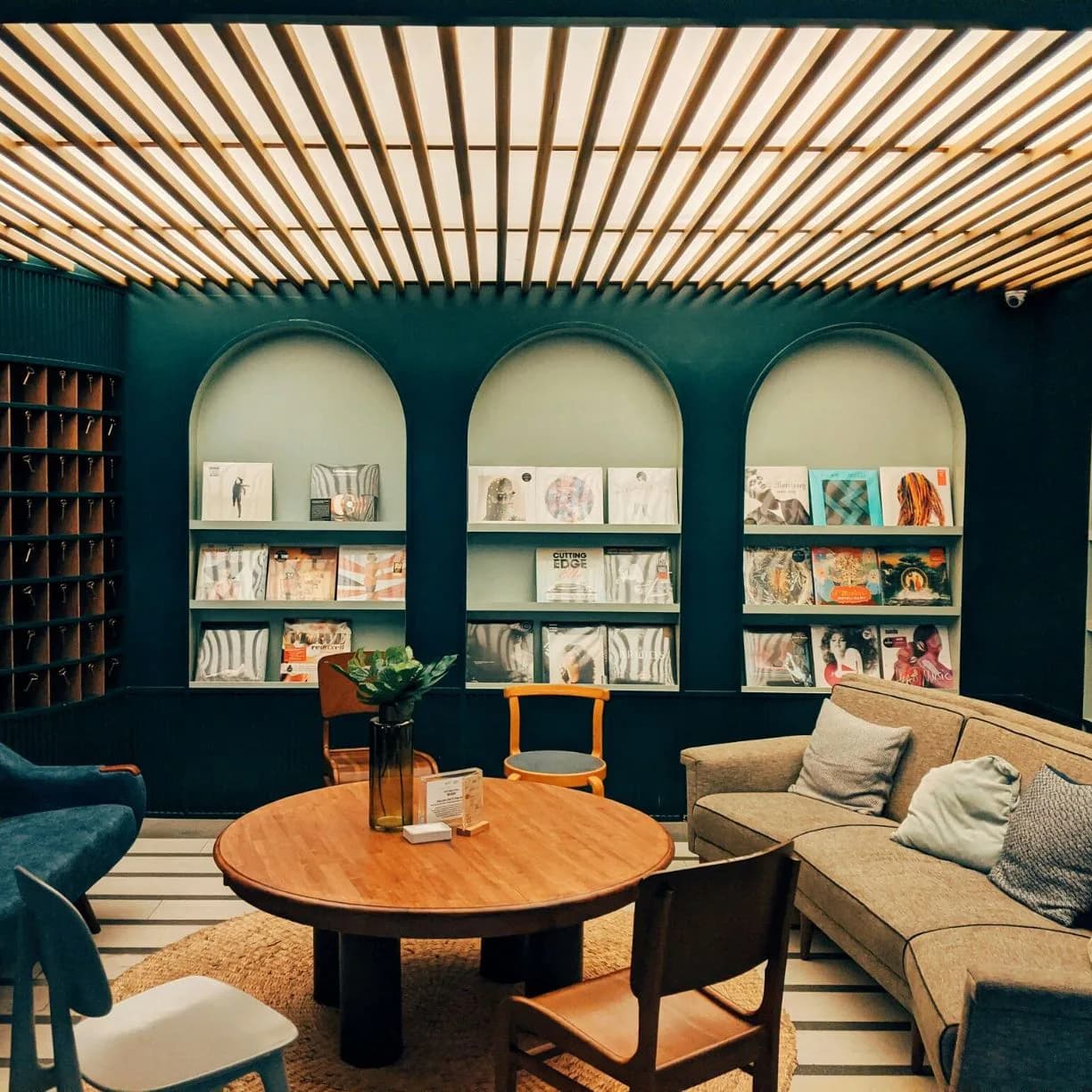 Teal-walled library lounge featuring three arched built-in shelving niches, round wood table, beige sectional sofa, and linear wooden ceiling by Juanita Jimenez.