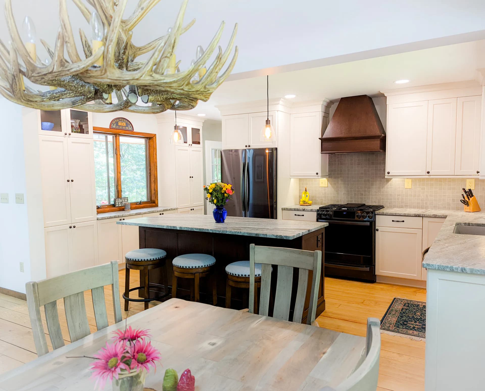 Bright modern kitchen with white cabinetry, black island with blue upholstered seating, stainless steel appliances, and distinctive antler chandelier, designed by Judi Price.