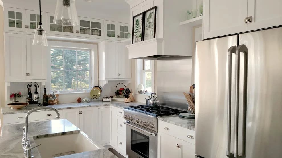 Bright white kitchen with stainless steel appliances, white cabinetry with glass uppers, marble waterfall island, and pendant lighting by Judi Price.
