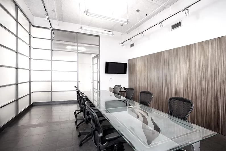 Modern office conference room with glass partition, wood-paneled accent wall, white glass table, black chairs, and mounted TV by Judith Gerrard.