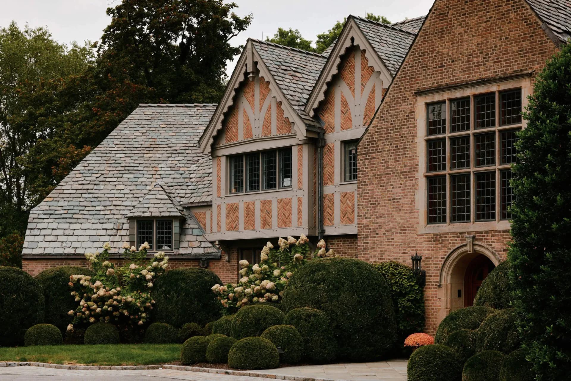 Tudor-style brick residence with steeply pitched roofs, decorative half-timbering, large casement windows, and manicured hedges by Judith Limon.