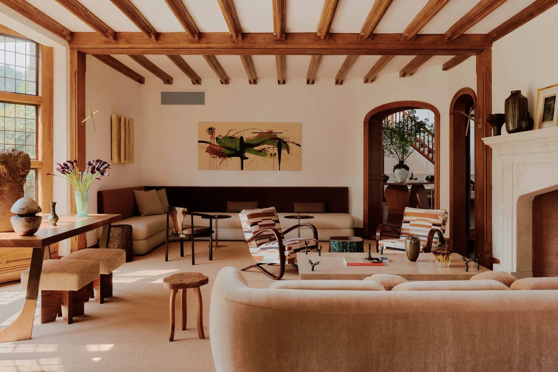 Warm-toned living room with exposed wooden ceiling beams, neutral upholstered seating, wooden furniture, and arched doorways, designed by Judith Limon.