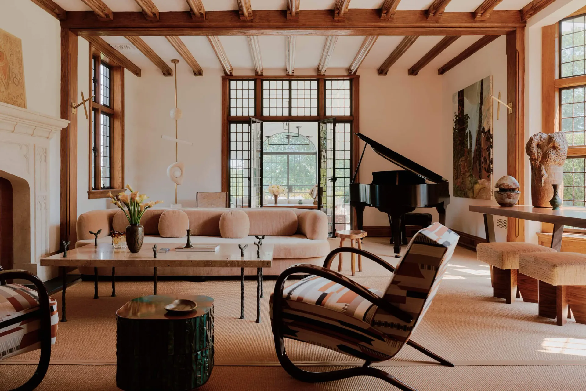 Modern living room with exposed wood beams, beige upholstered sofa, sculptural wooden chair, grand piano, and expansive windows overlooking water by Judith Limon.