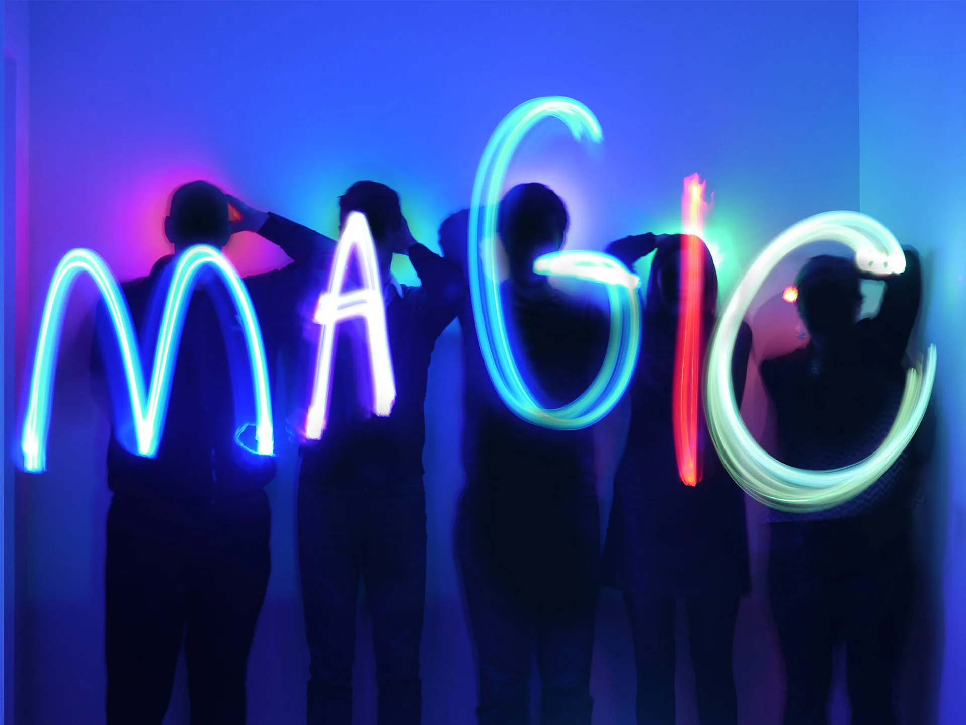 Neon light installation spelling 'MAGIC' in glowing blue, cyan, and pink tubes against a dark blue gradient backdrop, with silhouetted figures by Jules Fisher.