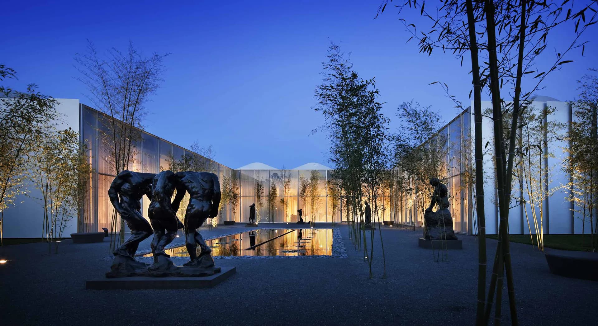 Twilight courtyard with illuminated modern pavilions, bronze elephant sculpture, and birch trees in an architectural installation by Jules Fisher.