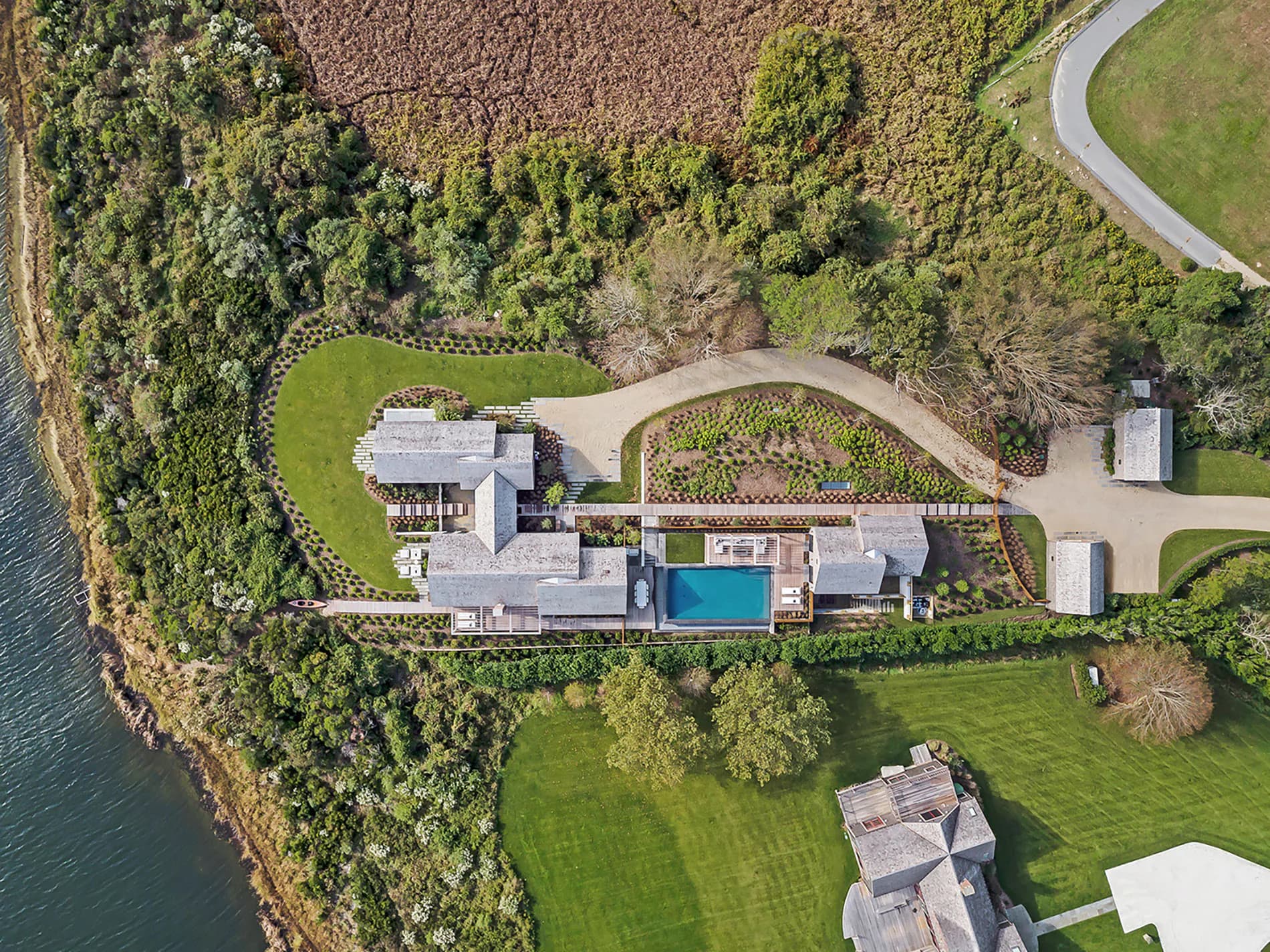Aerial view of a modern waterfront residence with clean lines, swimming pool, and manicured grounds nestled among dense green trees, designed by Julia Turner.