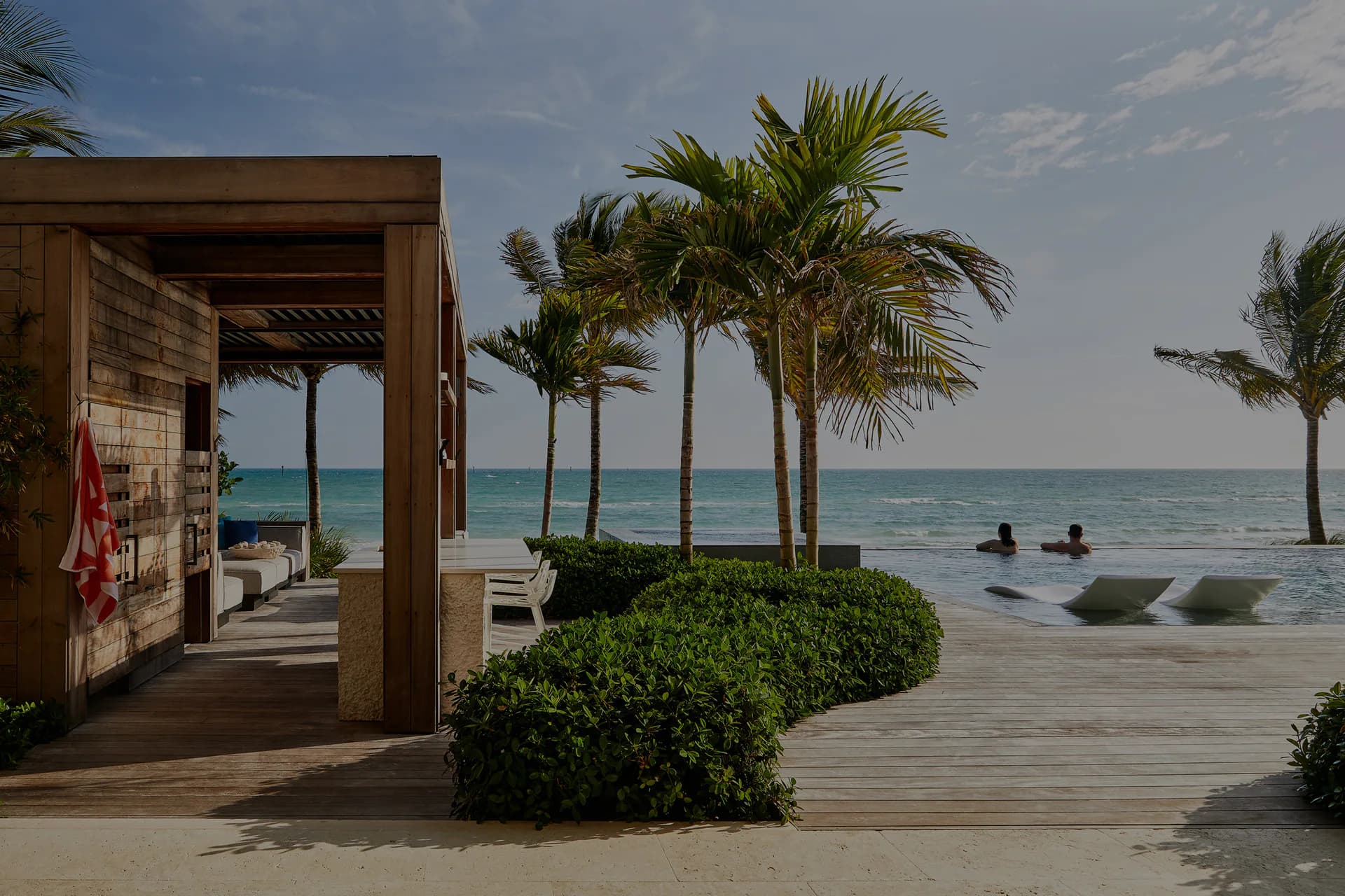 Coastal resort pavilion with wooden pergola, manicured hedges, and palm trees overlooking an infinity pool and ocean, designed by Julia Turner.