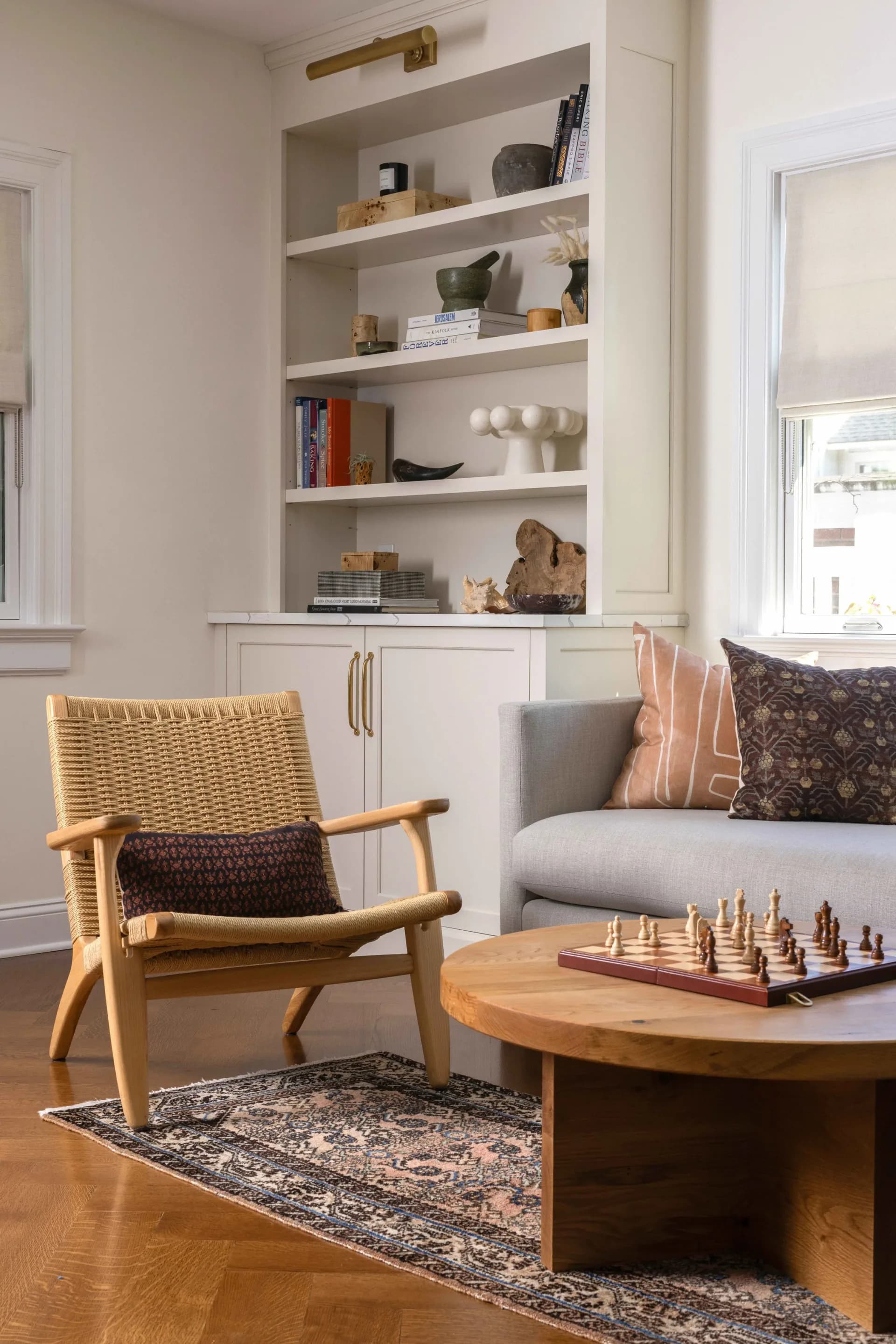 A bright, minimalist living room featuring a mid-century modern wooden chair with woven back, gray sofa, round wooden coffee table with chess set, and built-in shelving with decorative objects and books by Julieta Alvarez.